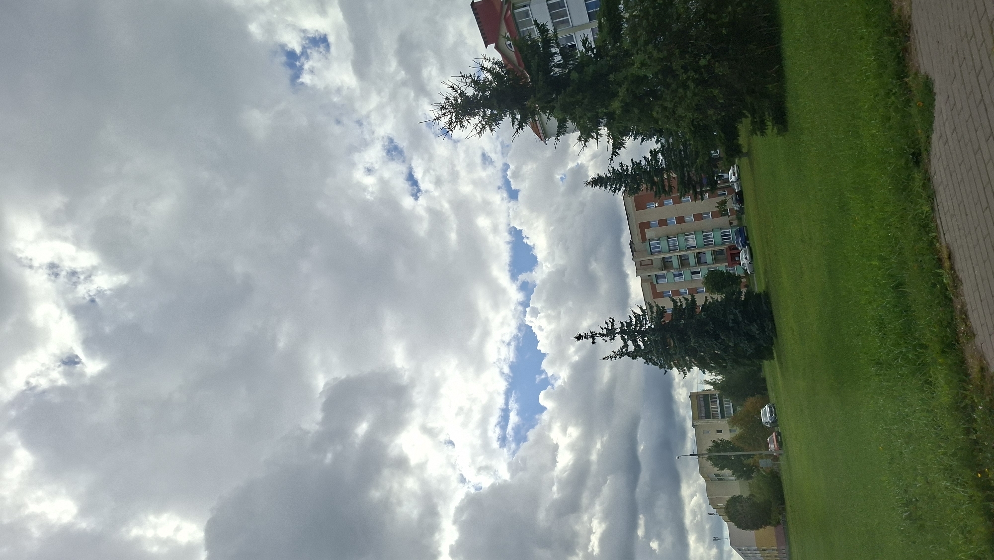 The image depicts an outdoor scene in Myadzyel, Belarus, with a focus on a cloudy sky, apartment buildings, and a grassy park area. The sky is filled with a mix of dark and light grey clouds, with patches of blue sky visible. In the middle ground, several multi-story apartment buildings are visible. The buildings are of varying architectural styles, with some appearing older and others more modern, featuring balconies and windows. To the right, a building with a red roof is prominent. In the foreground and middle ground, a well-maintained grassy area is visible, with several tall evergreen trees interspersed throughout. A paved walkway or sidewalk is visible in the lower right corner of the image. Several cars are parked on the street in front of the apartment buildings. The overall impression is a typical urban residential area in Belarus, under a dramatic sky. There are no people or animals visible in the image.