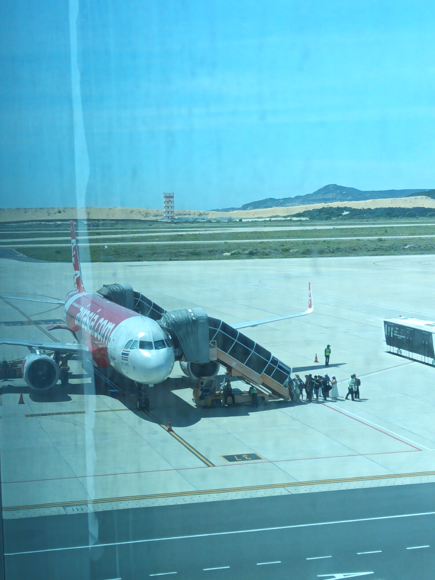 An AirAsia airplane is parked at an airport gate in Cam Ranh, Vietnam, viewed from inside a terminal building. The airplane, predominantly red and white, is connected to a jet bridge. Passengers are visible near the jet bridge, some disembarking and others waiting. A bus is parked to the right of the aircraft. The airport tarmac is visible, with runway markings and distant dunes and hills in the background under a clear blue sky.