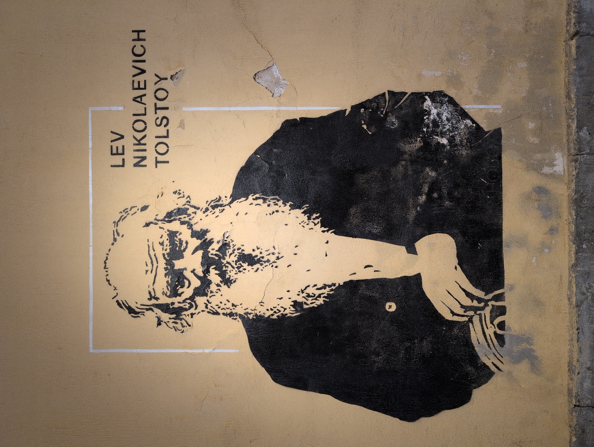 A stencil mural of Lev Nikolayevich Tolstoy is painted on a beige, textured wall. The stencil depicts Tolstoy from the chest up, facing slightly left, with his signature bushy beard and hair. He is wearing a dark jacket with a white collar visible. His left hand is resting on a book or papers. To the right of the portrait, the name "LEV NIKOLAEVICH TOLSTOY" is stenciled in black uppercase letters. The mural is framed by a thin, light-colored border. The wall shows signs of wear and tear, with cracks and peeling paint in several areas. The bottom of the image shows a concrete curb or foundation. The location of this mural in Saint Petersburg, Russia, is at the corner of Nevsky Prospect and Liteyny Prospekt, near the Fontanka River.
