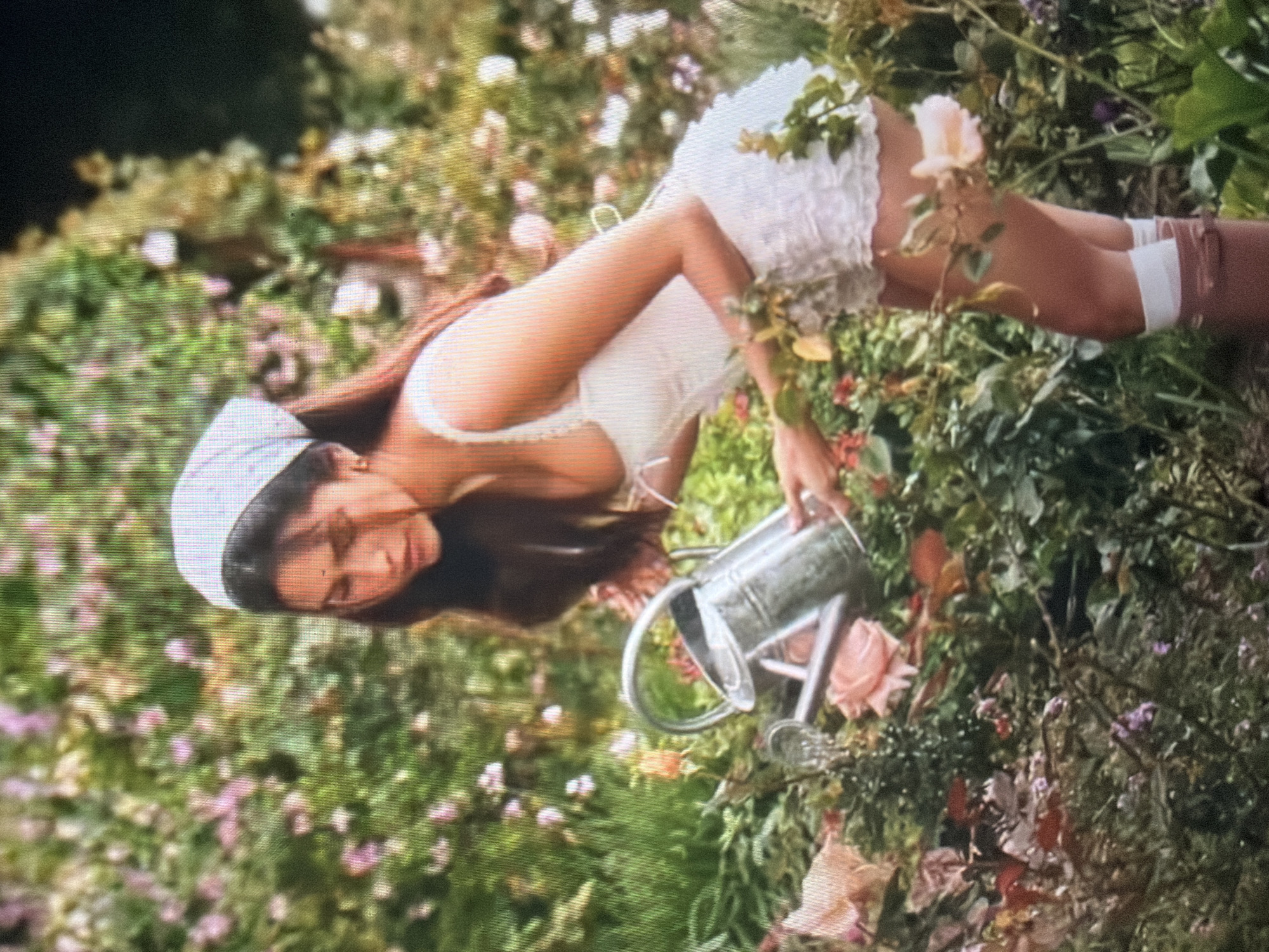 A young adult female is depicted in an outdoor garden setting. She has long, dark hair, partially covered by a white headscarf. A small dark mark is visible between her eyebrows. She is wearing a white corset-style top with thin shoulder straps and a short, ruffled white skirt, paired with white socks and what appear to be brown boots. The female is positioned bending forward, holding a silver-colored metal watering can with both hands, directing its spout towards a cluster of plants and flowers in front of her. The surrounding environment is a lush garden densely populated with various green plants and numerous blooming roses in shades of pink, peach, and white. The background foliage is blurred, indicating depth of field. The specific location in the city of Shumen, Bulgaria, cannot be determined from the visual information provided in the image.