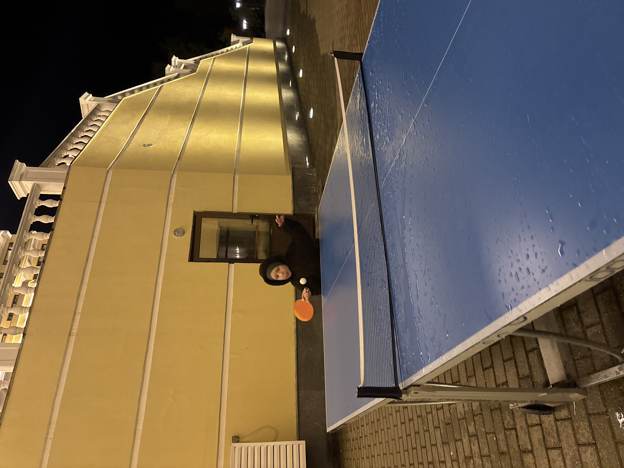 A person wearing a dark hooded jacket is playing table tennis at night. The person is holding an orange paddle and is in the process of hitting a white ball. The table tennis table is blue and appears to be outdoors on a brick-paved area. The table has a net across the middle, and the surface of the table is wet, possibly from rain. In the background, there is a large yellow building with white decorative elements along the roofline. The building is illuminated by yellow lights. There are also some small lights embedded in the ground in front of the building. The location is identified as Рождествено, Russia.