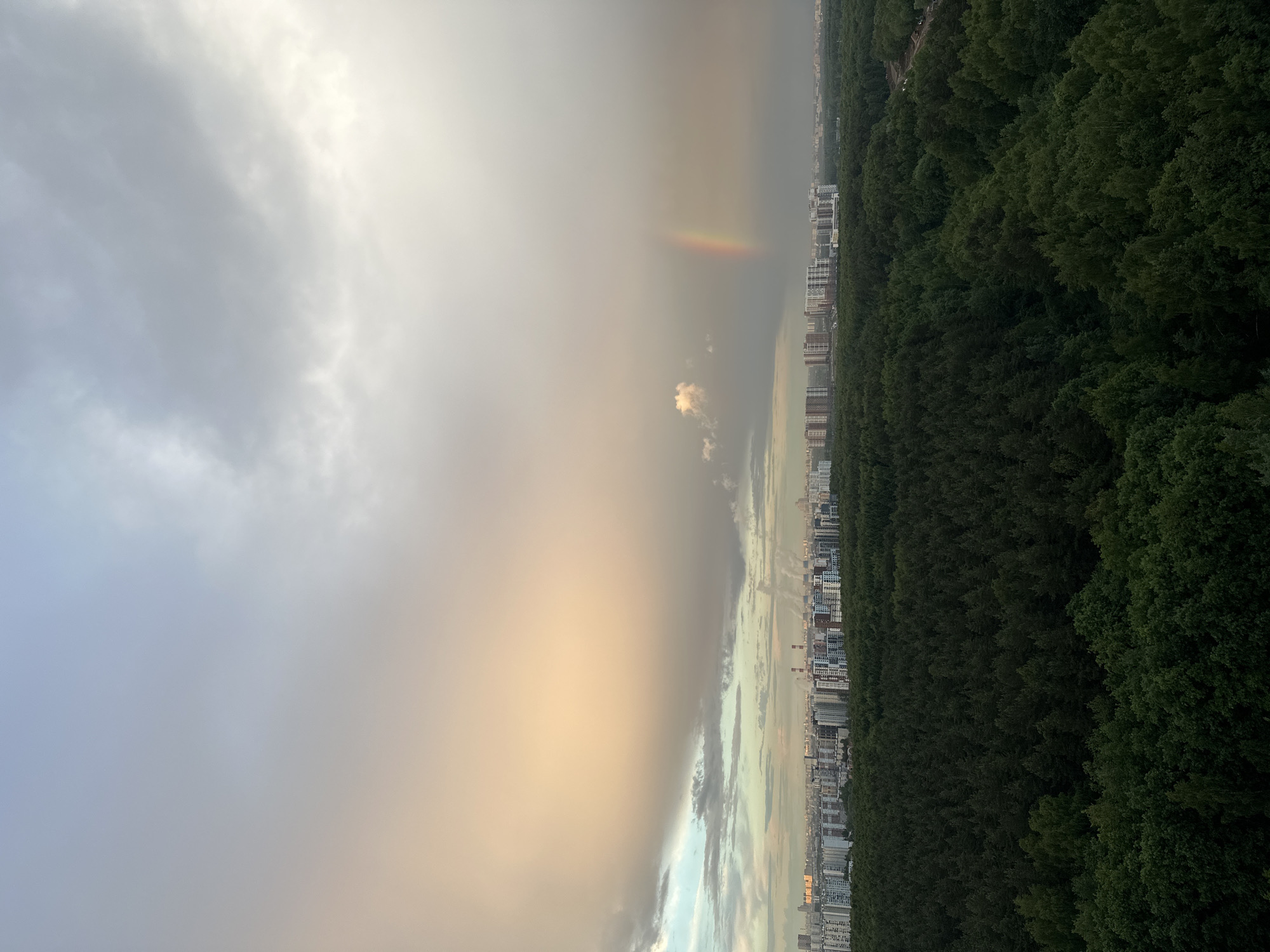An elevated landscape shot depicts a scene transitioning from cloudy weather to a partial rainbow and city buildings next to a green forest. The sky occupies most of the view, starting with heavy, gray clouds on the left that fade into a bright, cloudy area and reveal a section of a rainbow, indicating an atmospheric event with rain. To the right, the scene transitions into a cityscape made up of closely packed buildings that border a thick, green forest. There are no discernible people or actions observed in this shot.