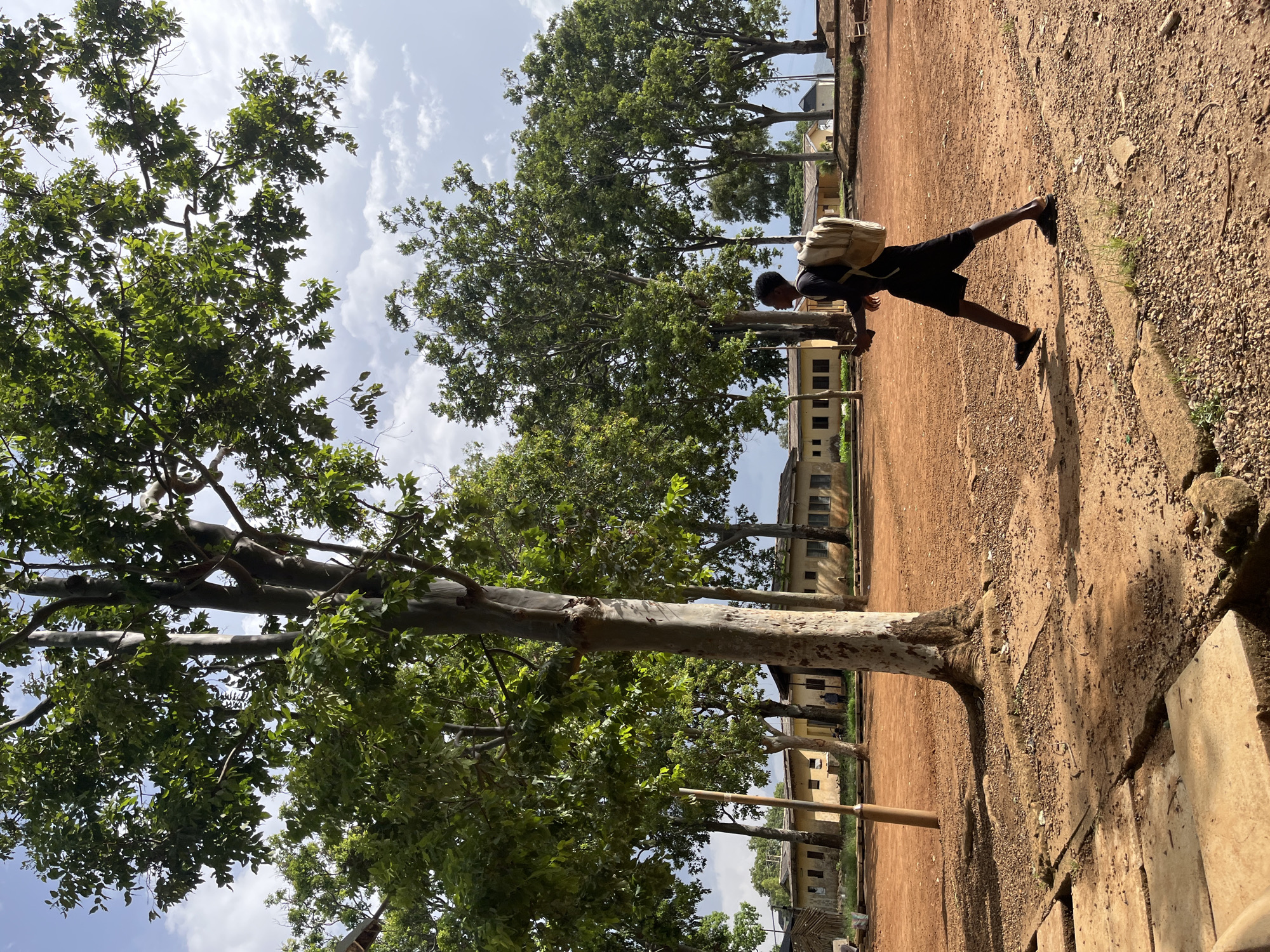 A young Black male wearing black shorts and a black t-shirt with a light-colored backpack walks across a dusty, reddish-brown dirt area. He is holding a dark-colored object in his right hand. He is walking from right to left in the frame, with his body angled slightly towards the viewer. In the background, a row of single-story buildings with light-colored walls and dark roofs are visible, suggesting an institutional setting like a school or administrative complex. Several trees with lush green foliage are scattered throughout the scene, with a large tree dominating the foreground on the left side of the frame, its trunk light gray and textured. The ground is uneven, with patches of gravel and concrete slabs. The sky is partly cloudy with a bright, hazy atmosphere, indicative of daytime. This scene appears to be an outdoor area within a city or town.