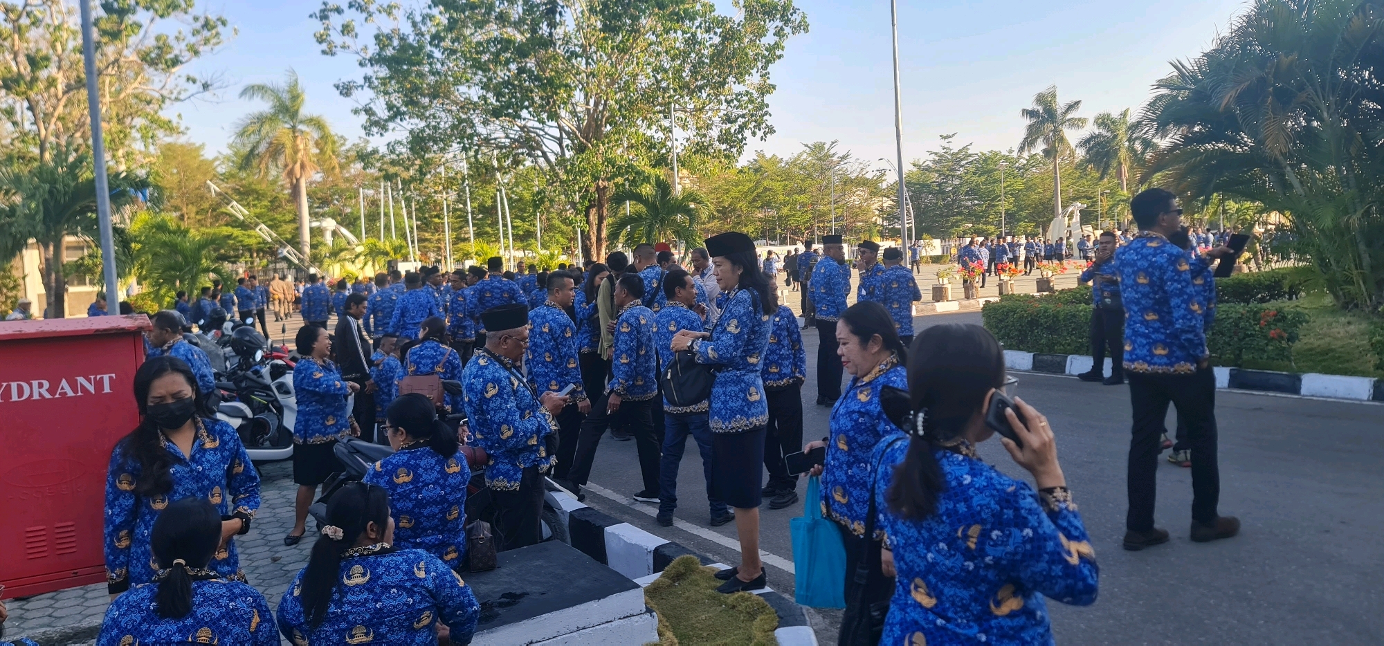 In Kupang, Indonesia, a group of individuals in matching blue batik shirts gather outdoors. The scene appears to be in a public space with a red hydrant labeled "HYDRANT" on the left. Many people are dressed in the same blue batik uniform, suggesting a formal event or gathering. Some individuals are talking amongst themselves, while others are looking at their phones. The environment includes trees and foliage, indicating a sunny day. Several motorcycles are parked in the background. The overall atmosphere suggests a community event or a workday gathering.