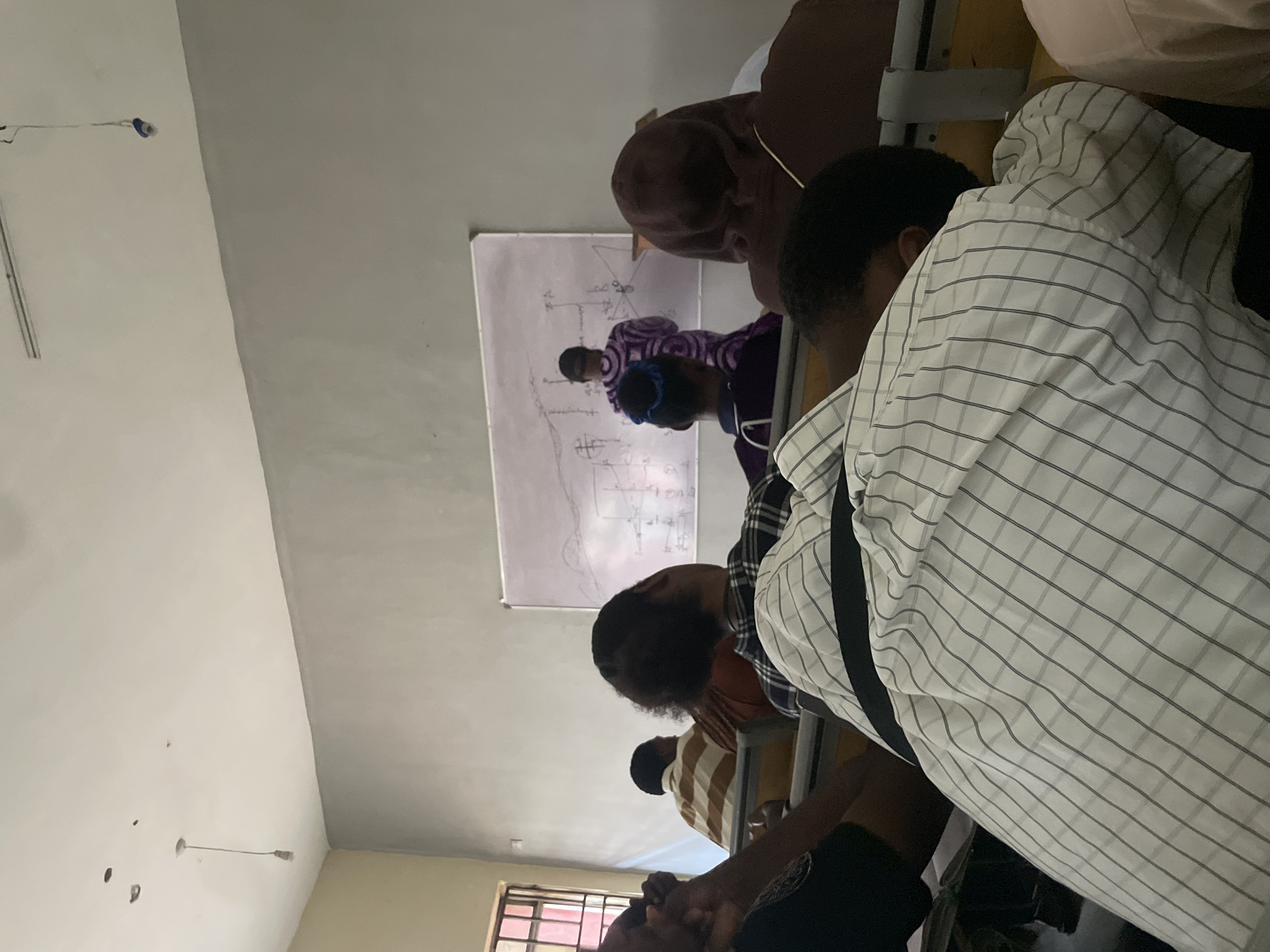 The image depicts a classroom scene in Ihiagwa, Nigeria, where a lecture is in progress. A lecturer, dressed in a purple patterned robe, stands at the front of the room, facing a whiteboard with diagrams and writing on it. Several students are seated in tiered rows, facing the lecturer. The foreground shows the backs of students, one wearing a white and grey checkered shirt. Another student is visible in a brown hoodie. The classroom has a grey wall on the right and a light-colored wall on the left. A window with barred panes is visible on the far left. The ceiling is plain and white, with a hanging light fixture. The overall atmosphere suggests an educational setting.