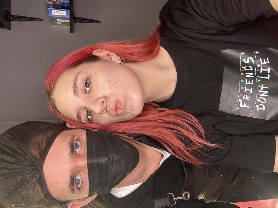 An indoor, eye-level photograph captures two individuals in what appears to be a hair salon or beauty establishment in Saratov, Russia. The individual on the left is an adult male, visible from the chest up. He wears a dark, disposable hairnet or cap, a black disposable face mask covering his nose and mouth, and a dark V-neck sweater over a white shirt. A dark, possibly leather, apron with metallic buckles is visible on his left shoulder and chest. His blue eyes are directed towards the camera. The individual on the right is a young adult female, also visible from the chest up. She has bright pink hair, styled long and straight. Her lips are pursed in a "kissing" gesture towards the camera, and her brown eyes are also directed forward. Acne or blemishes are visible on her right cheek. Her left ear displays two piercings: a hoop earring in the lobe and a stud in the helix. She wears a black crew-neck t-shirt featuring a graphic of string lights above the white text "FRIENDS DON'T LIE." The background consists of a plain, light gray wall. On the upper right, two dark, minimalist shelves are mounted, holding several small, rectangular blue and white bottles or boxes, possibly hair care products.