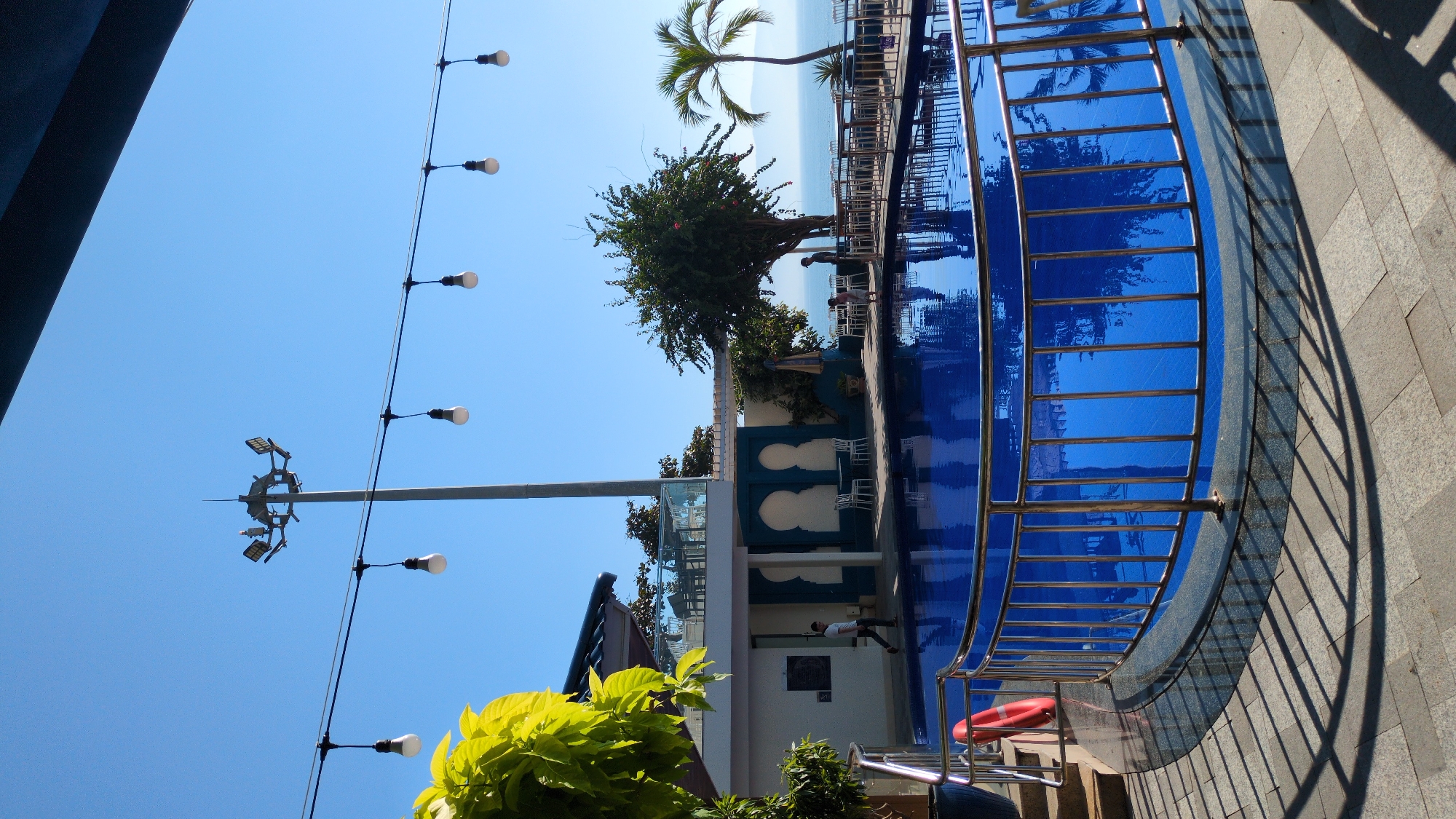 The scene depicts an outdoor leisure facility, likely a resort or hotel, situated in South Quy Nhon Ward, Vietnam, under a clear blue sky. A bright blue curvilinear swimming pool, partially enclosed by a polished metal railing, occupies the foreground and midground. A red lifebuoy is attached to the railing on the pool's left side. The surrounding pavement consists of light and dark grey square tiles, displaying distinct shadows cast by the railing.
To the left, a two-story building features white walls and a dark blue roof. The ground level presents two dark teal arched recesses with white outdoor tables and chairs positioned in front. A male individual, wearing a light-colored shirt and dark trousers, walks rightward across the paved area adjacent to this building. The upper level includes a balcony with glass railings and additional outdoor seating.
A tall grey utility pole, equipped with four visible floodlights and a pointed finial, rises prominently against the sky behind the building. String lights with white bulbs are suspended horizontally across the upper portion of the frame. To the right of the pool, on a lower patio area, an adult in dark attire stands beside a child in light clothing at the pool's edge, both facing towards the water. This area contains additional white tables and chairs, partially obscured by a striped umbrella.
The background features a large leafy green tree with visible red flowers and a slender palm tree. Beyond these, the blue ocean stretches to the horizon, with distant mountains or landmasses visible across the water.