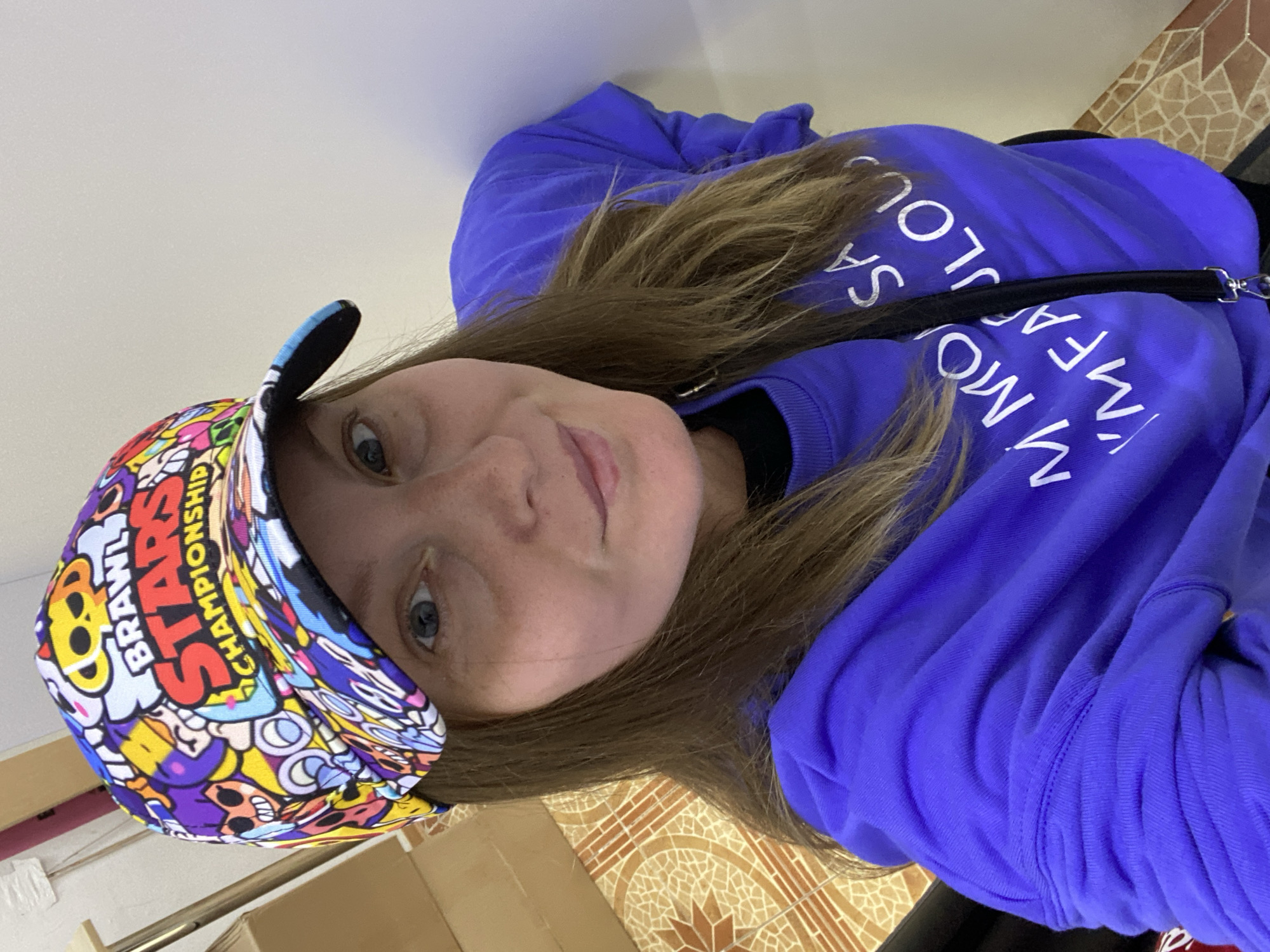 A woman wearing a purple sweatshirt and a colorful "Brawl Stars Championship" cap takes a selfie. The sweatshirt has white text that reads "I'M MORE FABULOUS". She has long brown hair and is looking directly at the camera with a slight smile. The background includes a tiled floor, a wall, and some cardboard boxes. The exact location in Noviy, Russia is not discernible from the image.