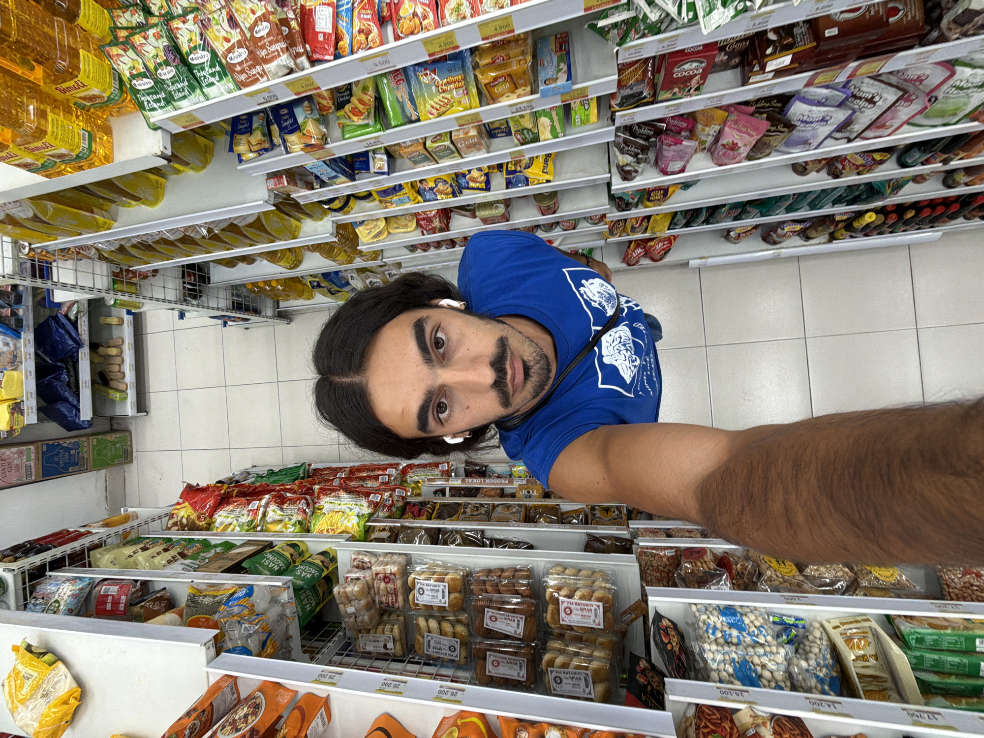 The image is a selfie taken by a man with long dark hair, a mustache, and a blue t-shirt in what appears to be a supermarket aisle in North Kuta, Indonesia. He is wearing wireless earbuds. The shelves are stocked with various food products, including cooking oil, snacks, and packaged meals. Many items have Indonesian text on their labels. Prices are visible on some items, for example 29.200 for Pia Baturiti and 9.500 for cheddar singles.