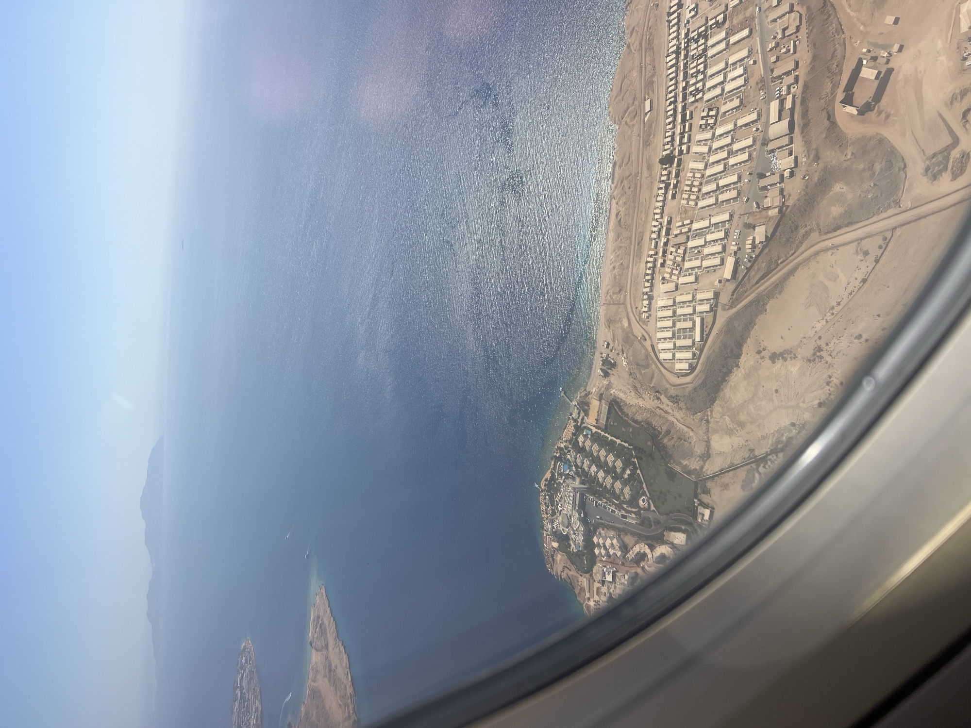 This is an aerial photograph taken from an airplane window, looking out over a coastal area. The predominant colors are the deep blue of the sea and the sandy beige of the land. In the distance, a chain of islands or mountains is visible on the horizon. The sea is calm with some light reflections on its surface.
The land in the foreground is arid and appears to be a desert landscape. Along the coastline, there is a developed resort area with numerous buildings, including a large complex with swimming pools and a marina or pier extending into the water. Further inland, there is a large, densely packed settlement consisting of many small, uniform buildings, possibly worker housing or a temporary settlement. Roads are visible winding through this settlement and connecting it to other areas. There are also some green areas, suggesting a golf course or landscaped areas within the resort. The overall impression is of a resort town situated in a desert environment.
The image is of Sharm El Sheikh, Egypt, a popular tourist destination known for its beaches, coral reefs, and desert landscapes. The resort area depicted likely represents one of the many hotels and complexes that line the coast, while the densely packed settlement may be related to the construction or operation of these facilities, or perhaps a separate community. The aerial perspective emphasizes the stark contrast between the developed coastal strip and the vast, barren desert beyond.