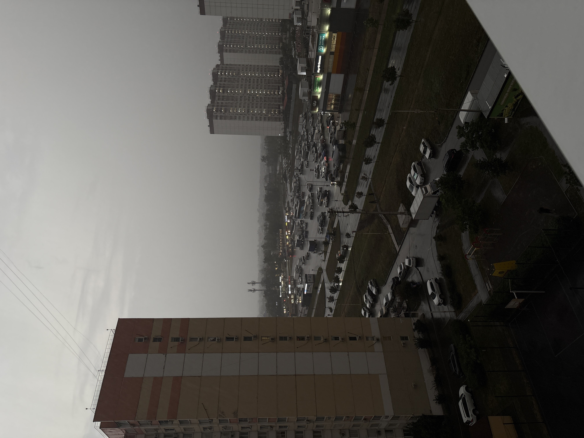 The image depicts an overcast, possibly rainy, urban scene. In the foreground to the left is a tall, multi-story residential building with a beige and orange facade. To the right of this building, a parking lot filled with numerous cars and trucks is visible. Beyond the parking lot, more tall residential buildings and commercial establishments with lit windows are present, suggesting a densely populated area. The ground appears wet, indicating recent or ongoing precipitation. A playground with yellow and red structures is visible in the lower right portion of the image, adjacent to a sports court. Electrical wires are strung across the sky in the upper left corner. The overall mood is somber due to the cloudy conditions. The presence of apartment buildings, a large parking area, and commercial storefronts suggests a residential and commercial district. Without further contextual information or visible landmarks, it is impossible to definitively confirm the location as Rostov-on-Don, Russia, or any other specific city.