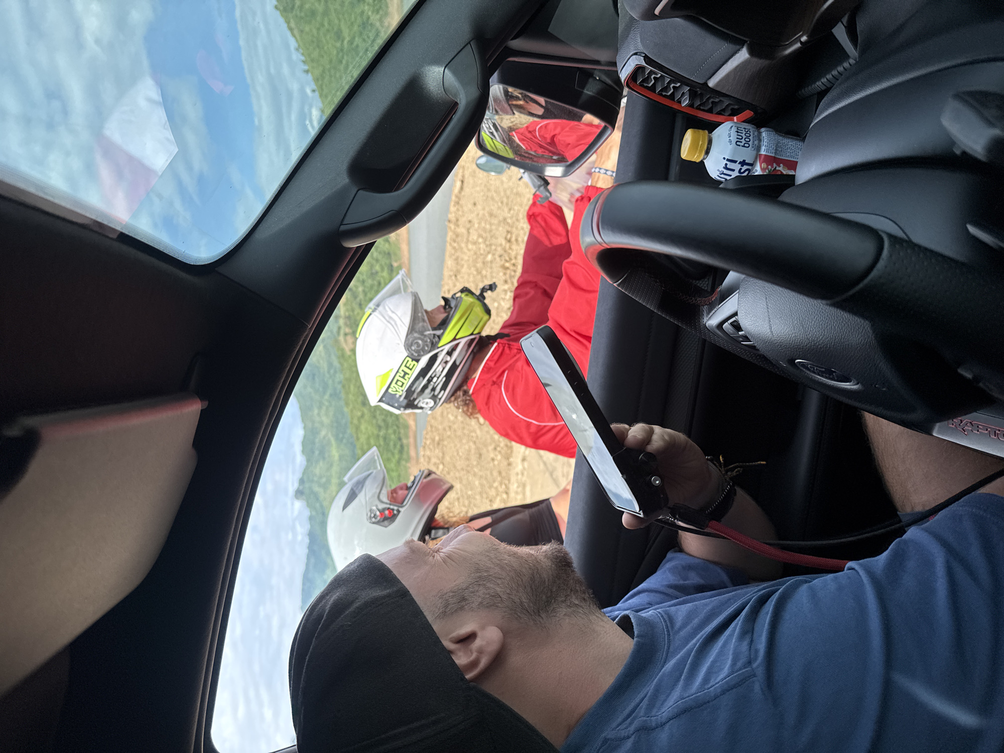 In Tân Phong, Vietnam, a man wearing a blue t-shirt and a black baseball cap is sitting inside a vehicle, holding a black device with a red cord attached. He is looking towards two individuals outside the vehicle. One individual is wearing a white helmet with a clear visor and a black shirt. The other individual is wearing a white and yellow helmet, a red jacket, and has curly hair. The background shows a body of water and lush green hills under a cloudy sky. The interior of the vehicle is visible, including the steering wheel, a bottle of "Nutri Boost," and the dashboard.