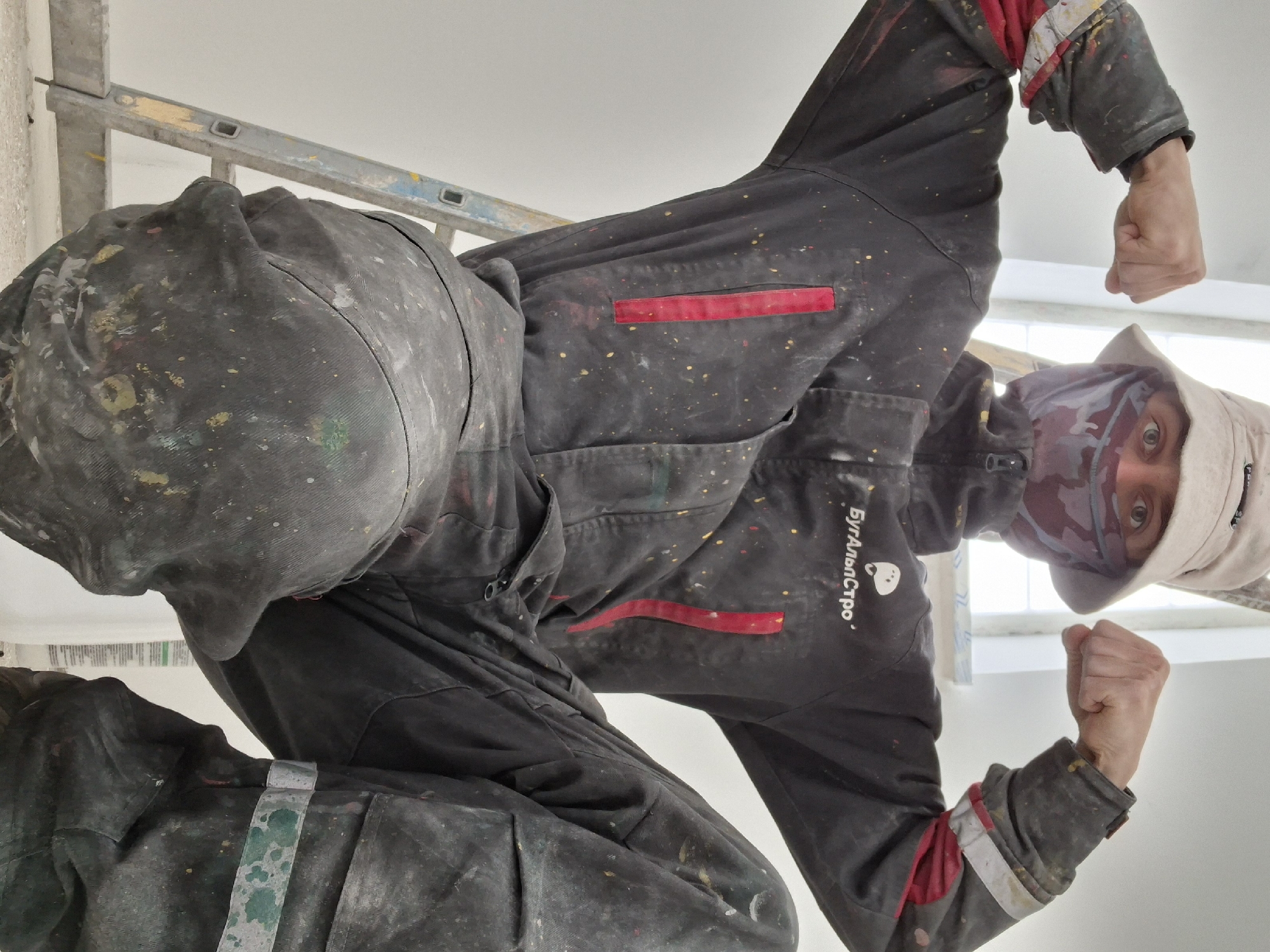 A painter, wearing paint-splattered black overalls, a beige hat, and a face covering, stands on a ladder in Unknown, Unknown. He holds his fists up, facing the camera, against a white wall and a window. The paint on his overalls is multicolored, and the ladder is metal. The text "БулАльпСтро" is visible on the overalls.