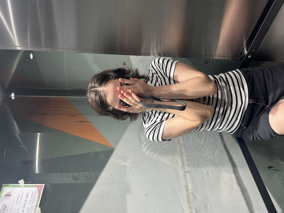 A person in a striped shirt and shorts stands in what appears to be an elevator. They have their hands covering their face, possibly out of embarrassment or discomfort. A camera strap hangs around their neck. The elevator walls are metallic and have geometric designs on the bottom half. A sign is affixed to the bottom left corner of the elevator interior.