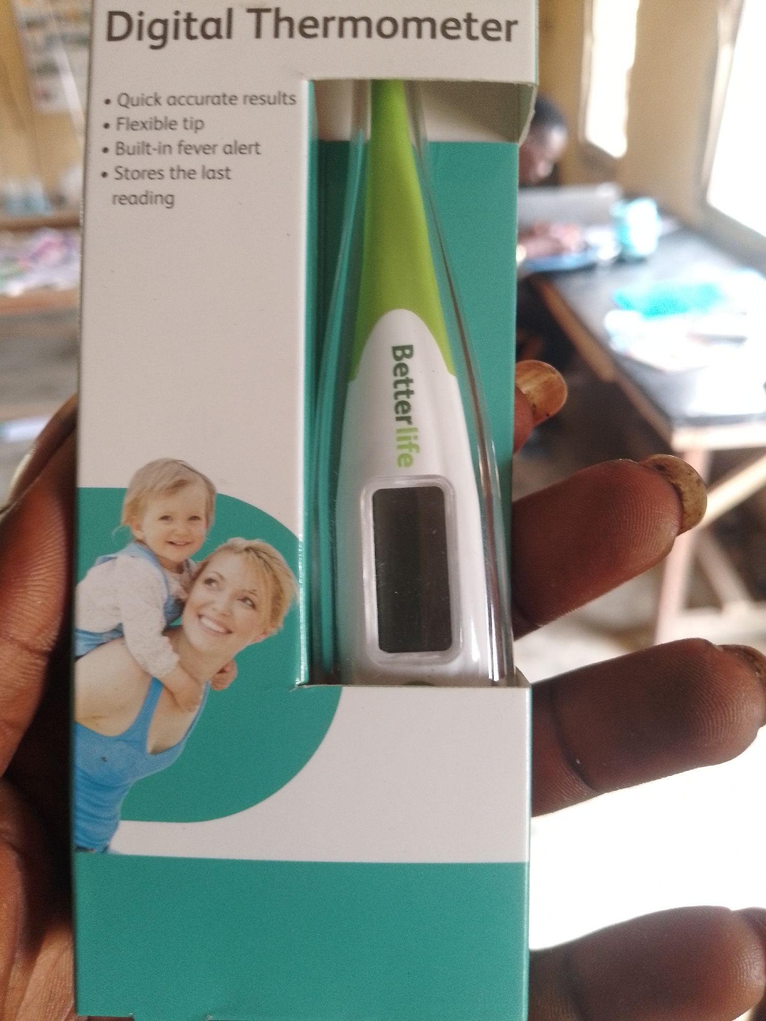 The image shows a close-up of a person's hand holding a white and green box for a "Digital Thermometer." The box has text listing features like "Quick accurate results," "Flexible tip," "Built-in fever alert," and "Stores the last reading." A graphic on the box depicts a woman carrying a smiling baby on her shoulders. A digital thermometer with the brand name "Betterlife" is partially visible within the box opening. The background is blurred, showing what appears to be an indoor setting with a table and a person in the distance. The photograph is taken in a candid, home-like environment.
