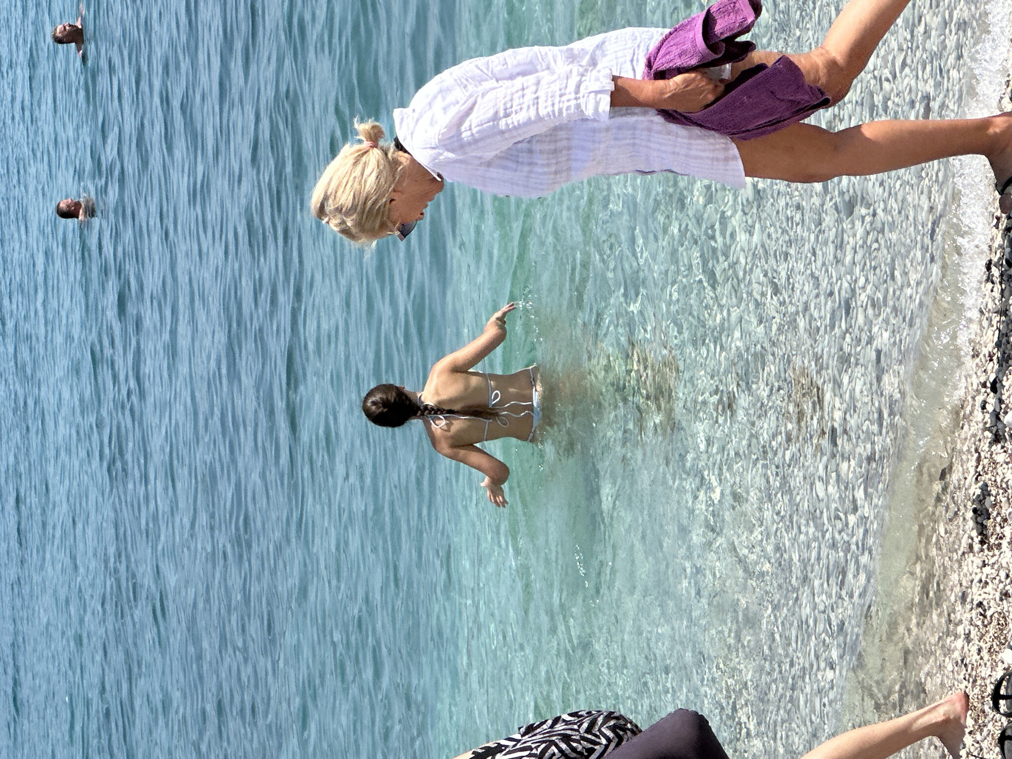 The image depicts a sunny day at a beach in Budva, Montenegro. Several people are visible in the clear, shallow turquoise water. In the foreground, a person's legs are visible as they sit on the pebbly shore. Further out in the water, a woman in a white shirt and purple shorts stands, holding a towel. To her right, a woman in a bikini is partially submerged in the water, facing away from the camera. In the background, two heads are visible above the water, suggesting they are swimming or wading. The water is calm and appears to be quite shallow near the shore. The overall impression is of a relaxing summer day at the beach.