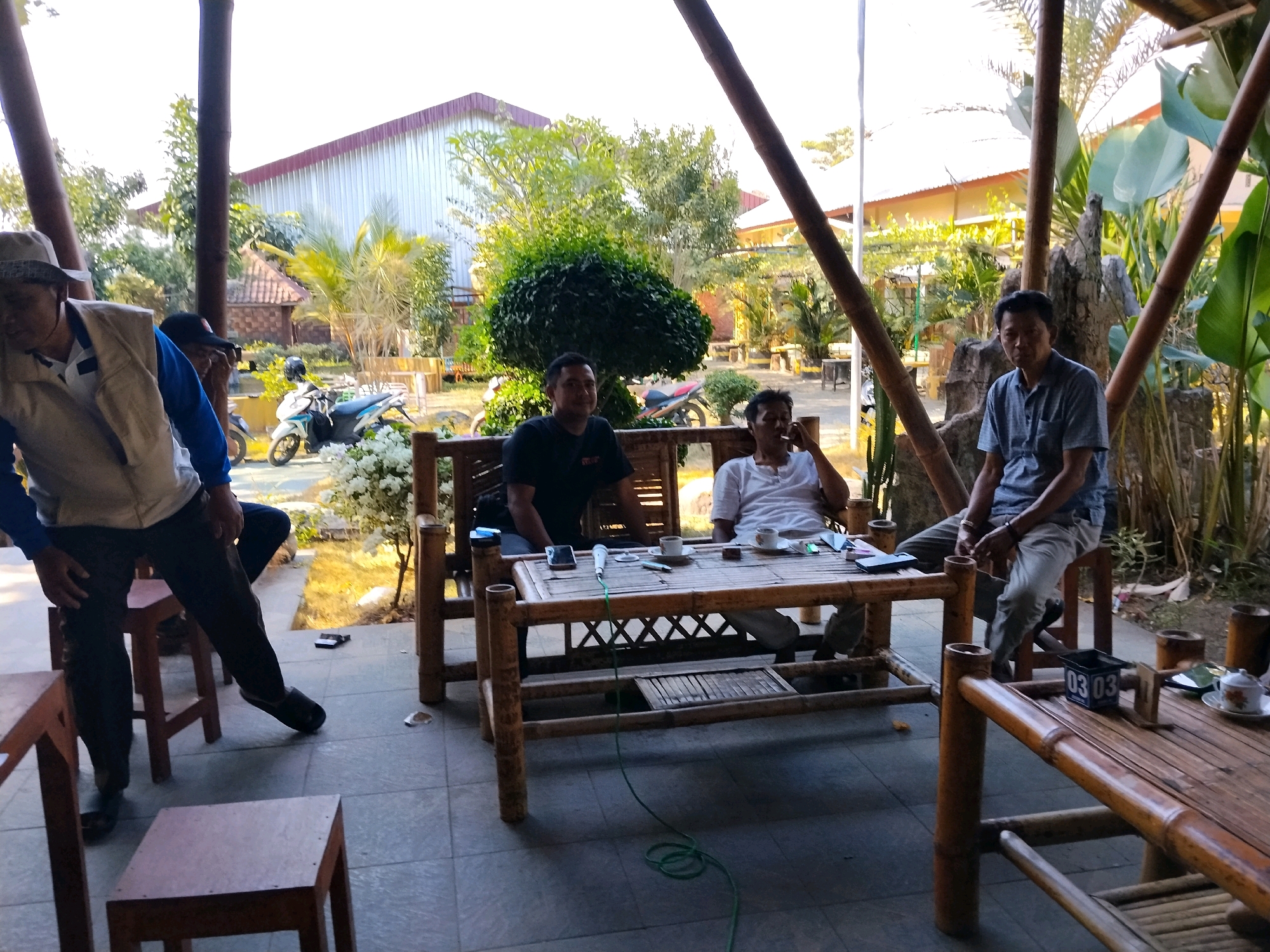 In a shaded outdoor area, likely a cafe or restaurant in Pekalongan, Indonesia, four men are gathered. Two men sit on a bamboo bench, one wearing a black t-shirt and the other a white t-shirt, both facing a bamboo table. The man in the white t-shirt is smoking. On the table are two cups of coffee, a mobile phone, a microphone, and other items. To the right, another man in a blue shirt and grey pants sits on a stool facing the table, holding what appears to be a small black box with numbers on it. To the left, a man wearing a hat, vest, and blue shirt is partially visible, appearing to be in motion. In the background, there are more bamboo structures, lush greenery, and buildings with red roofs. Motorbikes are parked in the distance. The ground is tiled. The lighting suggests it is daytime.