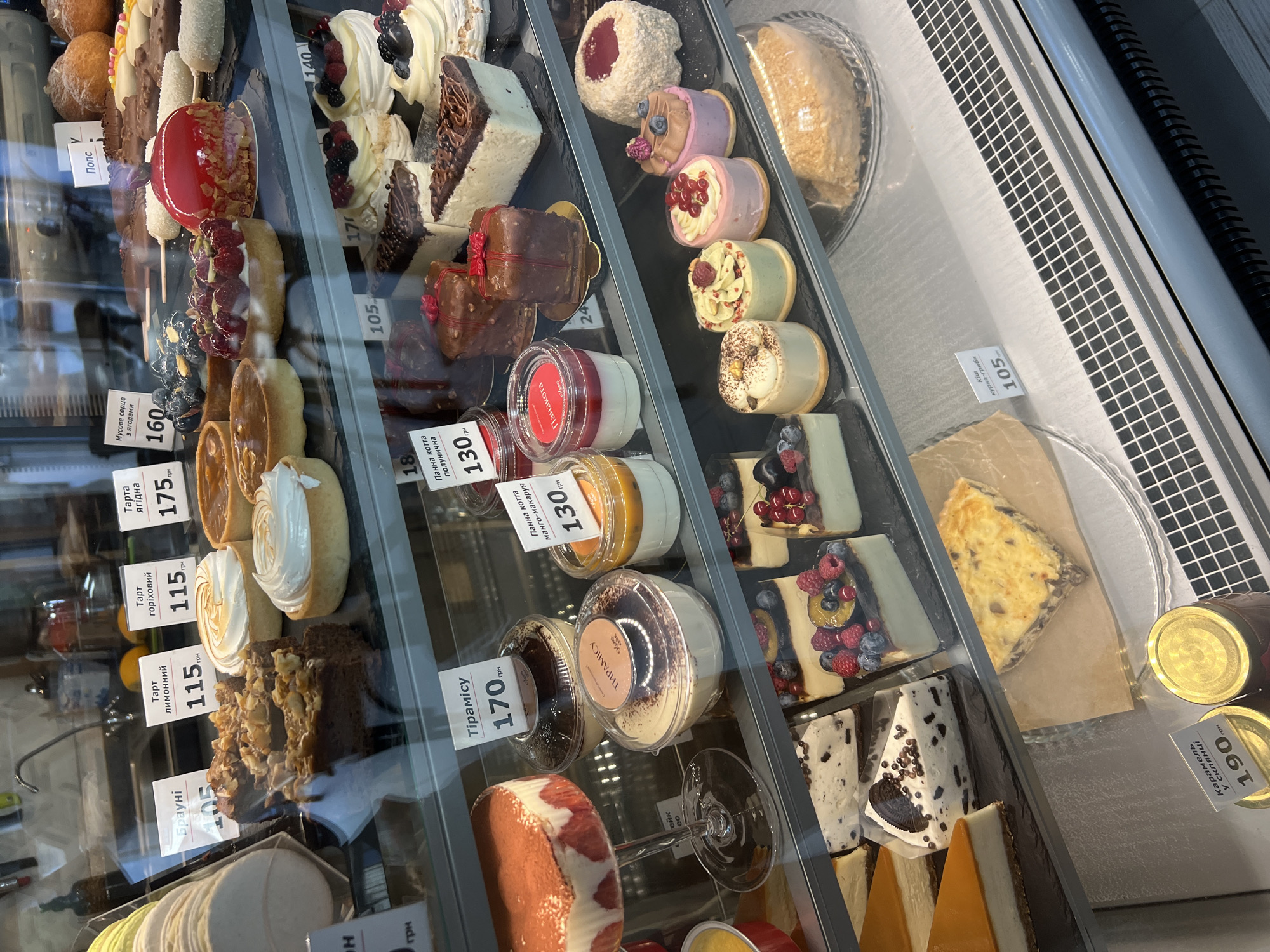 In a bakery display case, an assortment of pastries and cakes are arranged on multiple shelves. The items include tarts with fruit and cream toppings, layered cakes with chocolate and fruit decorations, individual desserts in plastic containers, and larger cakes, some partially visible. Price tags in Ukrainian currency are visible next to many of the items. The overall environment appears to be a commercial food establishment, likely a bakery or patisserie, located in Fontanka, Ukraine. There are no people visible in the frame, and no actions or interactions are taking place. The lighting is bright and focused on the display.