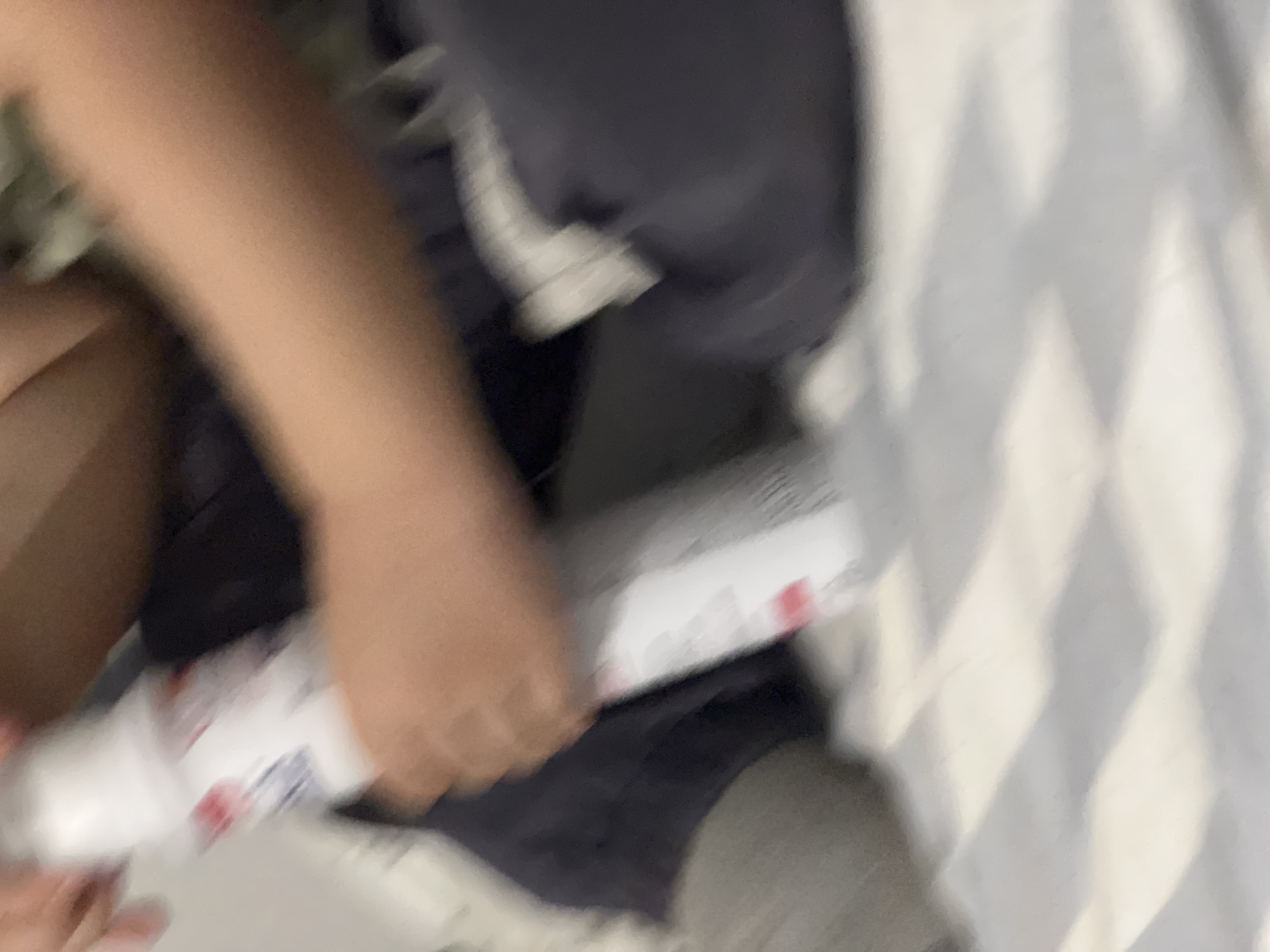 An out-of-focus, close-up shot shows a person's hands holding a white bottle with red and blue markings. The background is blurry and appears to be a bed with striped bedding. The scene is indoors.