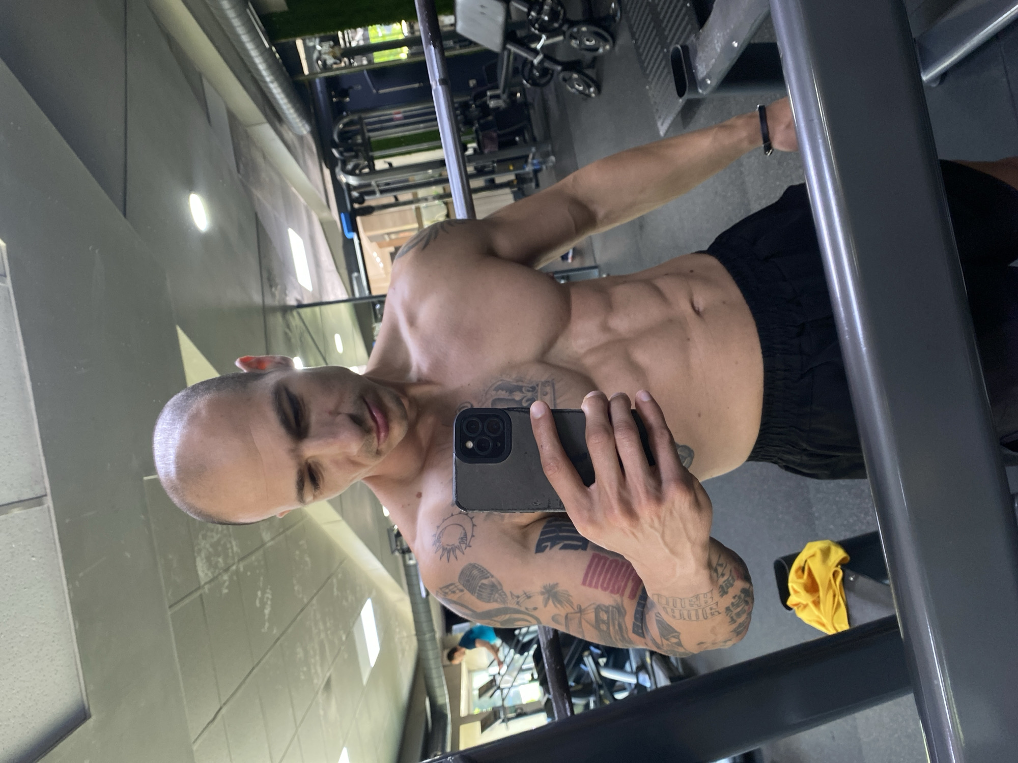 An image, rotated 90 degrees clockwise, captures a shirtless man taking a mirror selfie in a gym setting. The man, who appears to be bald and muscular, is positioned centrally in the frame. He has numerous tattoos covering his arms, shoulders, and chest, including what resembles a sun or moon on his left shoulder, a microphone, palm trees, and various other designs. One tattoo on his right forearm has red and white stripes. He is wearing black athletic shorts and a dark-colored wristband on his right arm.
He holds a dark-colored smartphone with a triple camera array in his left hand, angling it towards the mirror to capture his reflection. His gaze is directed towards the camera. The background reflects the interior of a gym, featuring various pieces of exercise equipment with metal frames and weights. The walls are light-colored, possibly gray or beige, with rectangular light fixtures visible. In the reflection further back, another person wearing a blue shirt can be seen. A yellow piece of cloth or towel is tucked into a piece of gym equipment near the man's right side. The overall lighting is bright, typical of an indoor fitness facility. The setting is a gym in Pleven, Bulgaria. No explicit text is clearly legible on the tattoos or objects.