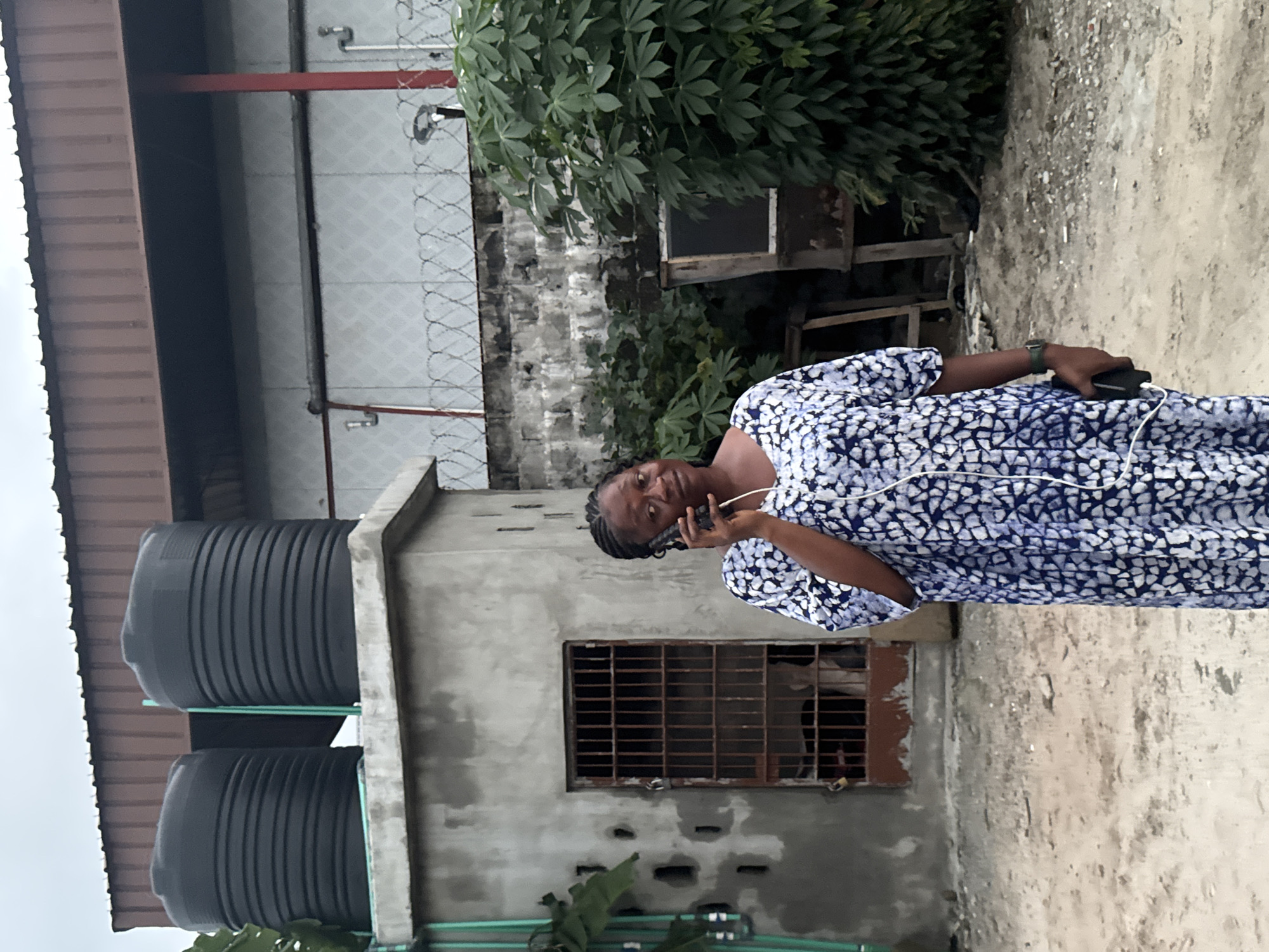 A Black woman stands outdoors, wearing a blue and white patterned dress and holding a smartphone to her ear. She has her hair braided and is wearing a watch on her left wrist. Behind her is a grey concrete structure with a barred window. Two large black water tanks are visible on top of the structure. The background features a building with a corrugated metal roof, a concrete wall topped with barbed wire, and lush green foliage. The ground is covered in dirt and gravel. The scene appears to be in a semi-industrial or residential area, possibly in Ajara, Nigeria.