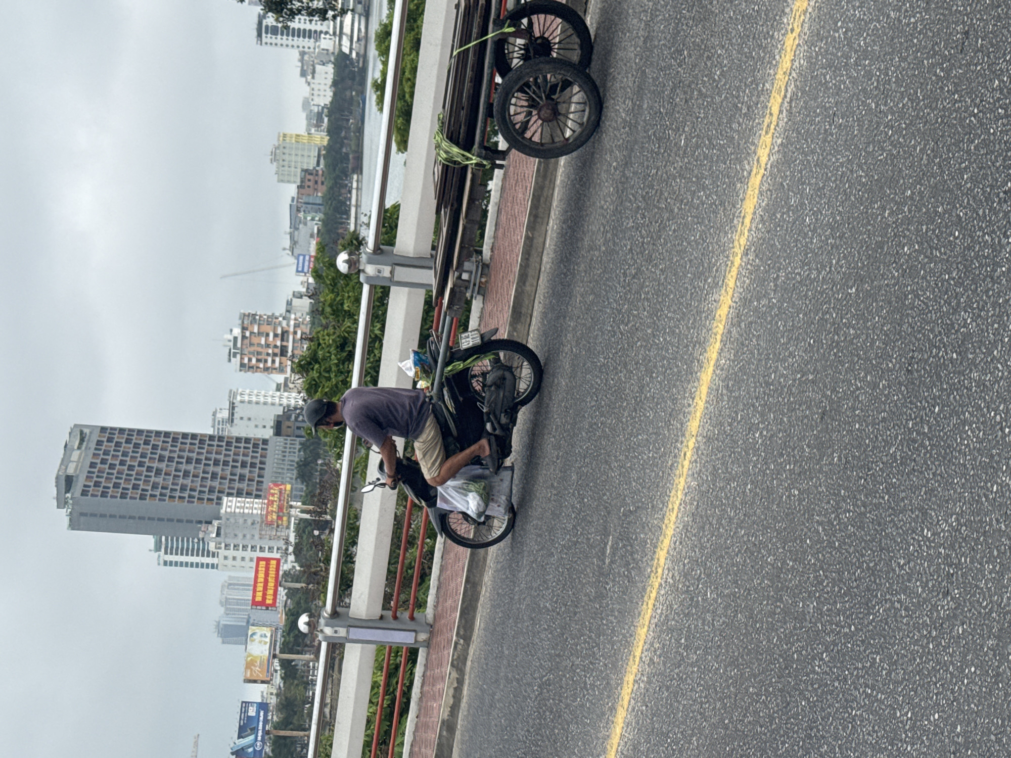 A man riding a motorcycle is positioned on the right side of a city street in Da Nang, Vietnam, near the railing of a bridge. He is wearing a dark shirt, light shorts, and a dark cap. The motorcycle is black, and additional items are loaded on it. Adjacent to him, near the railing, is a two-wheeled cart with items strapped on top. The street has a yellow line marking a lane. In the background are buildings, including a tall hotel and several smaller structures, visible through the overcast sky.