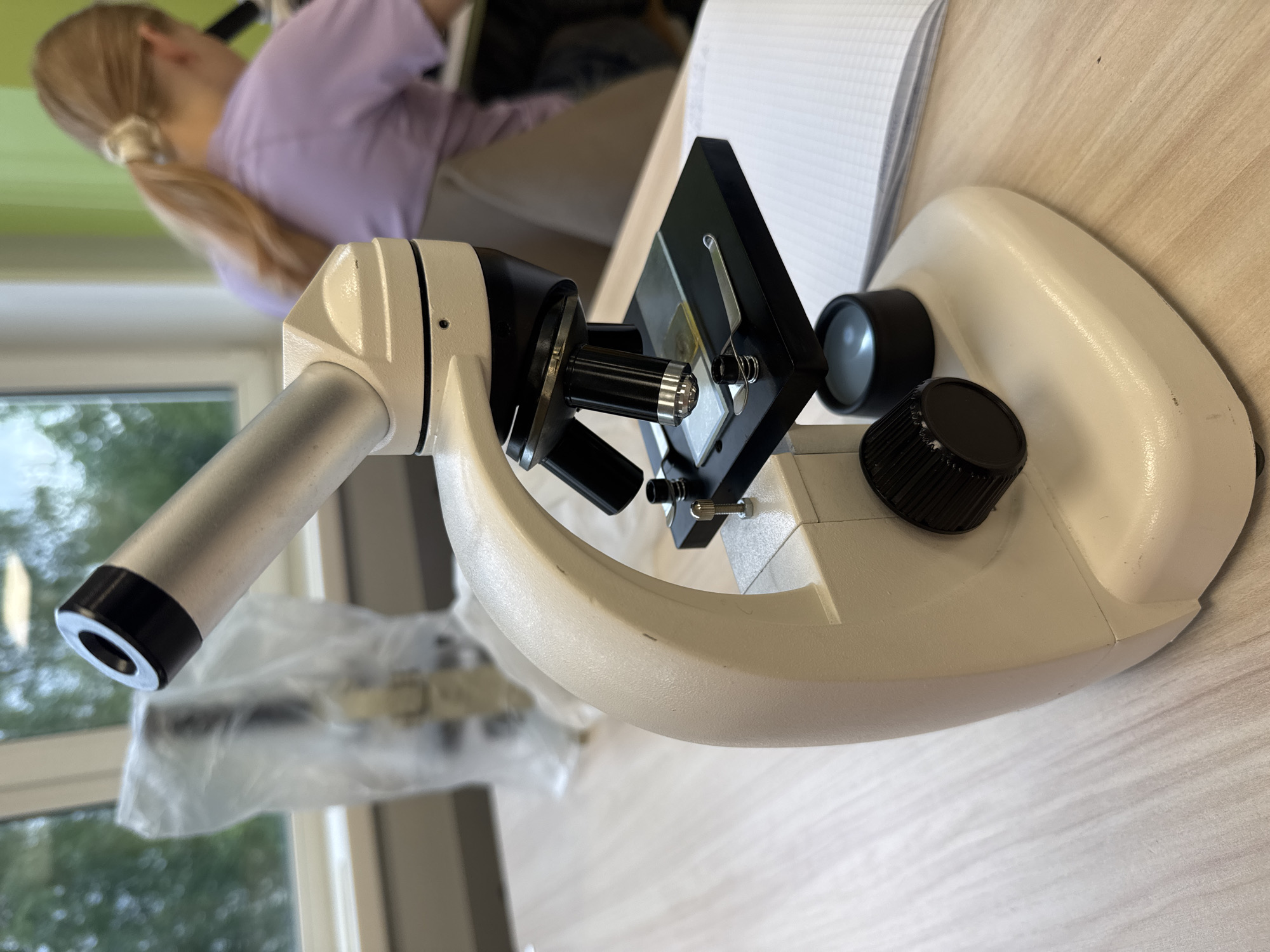 The image shows a white microscope on a light-colored wooden surface. The microscope's objective lenses are visible. A black stage with a slide clip holds a microscope slide. A black knob is located on the right side of the microscope base. In the background, a person with blonde hair pulled back into a ponytail and wearing a light purple shirt is visible, sitting at what appears to be a desk with papers. A window with a view of trees is in the background. Given the detail, this image may have been captured during a microscopy lesson or in a laboratory. There is no evidence present in this image that could connect the location of this photo to the city of Chaikovsky, Russia.
