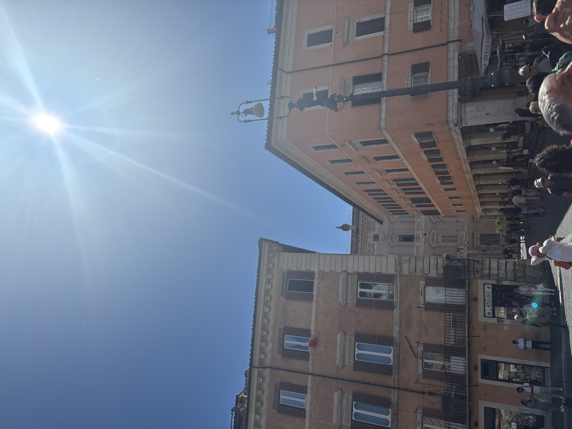 The image depicts a street in Rome, Italy, viewed from a low angle with the buildings dominating the frame. The sky is bright blue with a glaring sun. On either side of the street, there are tall buildings of similar height and architectural style, with multiple stories, rows of windows with shutters, and a pale peach or tan color. A street light is visible, suspended above the street between the buildings. The street is populated with people, many of whom are blurred, suggesting movement. A shopfront is discernible on the building on the right side of the frame.