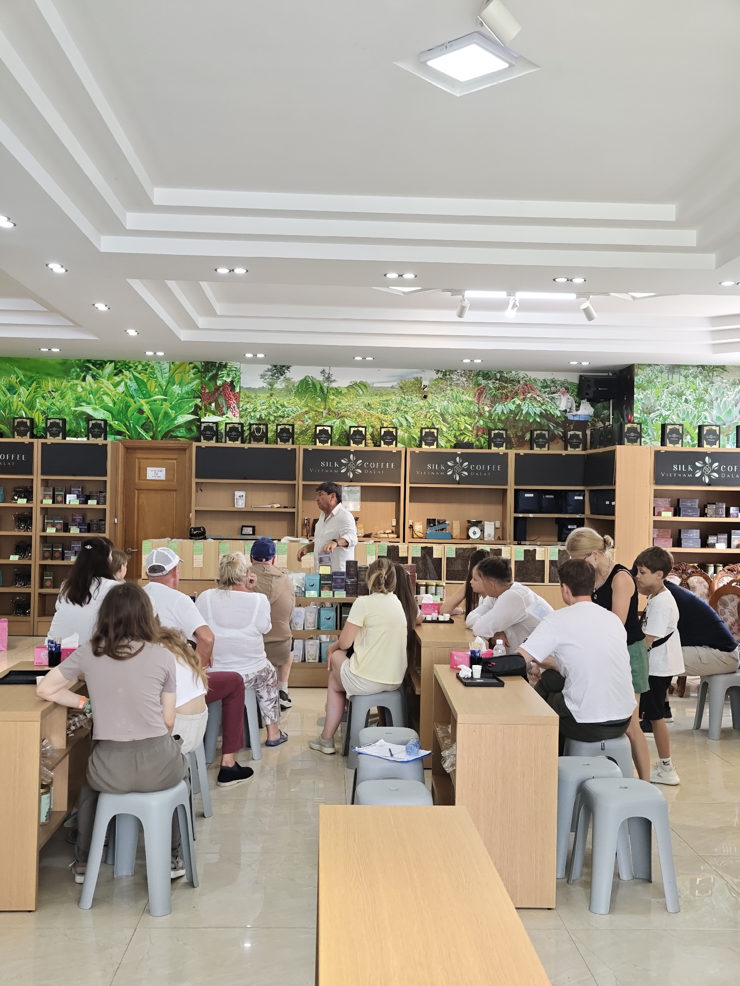 The image depicts an indoor scene within a shop in Lam Vien - Da Lat, Vietnam. The shop appears to be a specialty store selling coffee and silk products, suggested by signage visible on the shelves: "SILK COFFEE VIETNAM DALAT." The environment is well-lit with modern recessed lighting and track lighting in the ceiling, which features a stepped design.
A group of approximately 15-20 people, appearing to be tourists and possibly staff, are gathered. Most are seated on light grey plastic stools or chairs at wooden counters. Several individuals are standing, including a man in a white shirt who appears to be addressing the seated group, gesturing with his hands and holding a small object. The seated individuals are facing forward, some looking towards the speaker, while others are looking down or at their hands.
The shop's interior is characterized by wooden shelving units filled with merchandise, including boxes and bags of coffee and other products. A large mural of lush green tea plants and coffee beans adorns the wall behind the shelves, providing a vibrant backdrop. The floor is tiled with a light-colored, reflective surface. The overall atmosphere suggests a presentation, tasting, or educational session about the products being sold.