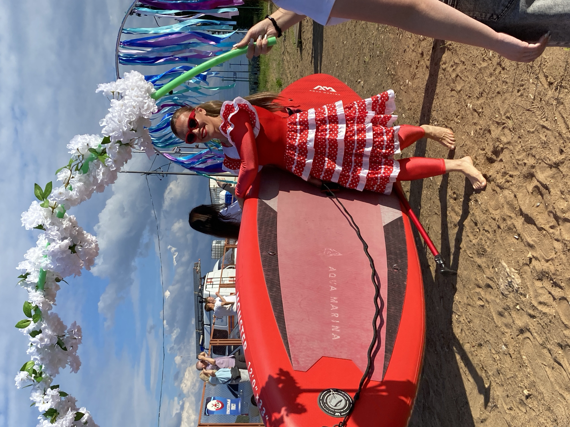 In Chaikovsky, Russia, a woman in a vibrant red and white polka dot dress, adorned with ruffles and red sunglasses, poses playfully on a red paddleboard. She is holding a green balloon on a stick. The paddleboard is resting on a sandy ground, possibly near a body of water, given the presence of other people in casual attire and what appears to be a pier or boat dock in the background. A large floral archway, constructed from white artificial flowers, frames the scene on the left, with blue and purple ribbons fluttering in the background. The sky is bright blue with scattered white clouds, indicating a sunny day. The overall mood is festive and lighthearted, suggesting a celebration or a special event. The text "AQUA MARINA" is visible on the paddleboard, along with "MONSTER 12'0" X 33"". There is also a sign with a red cross and Russian text.