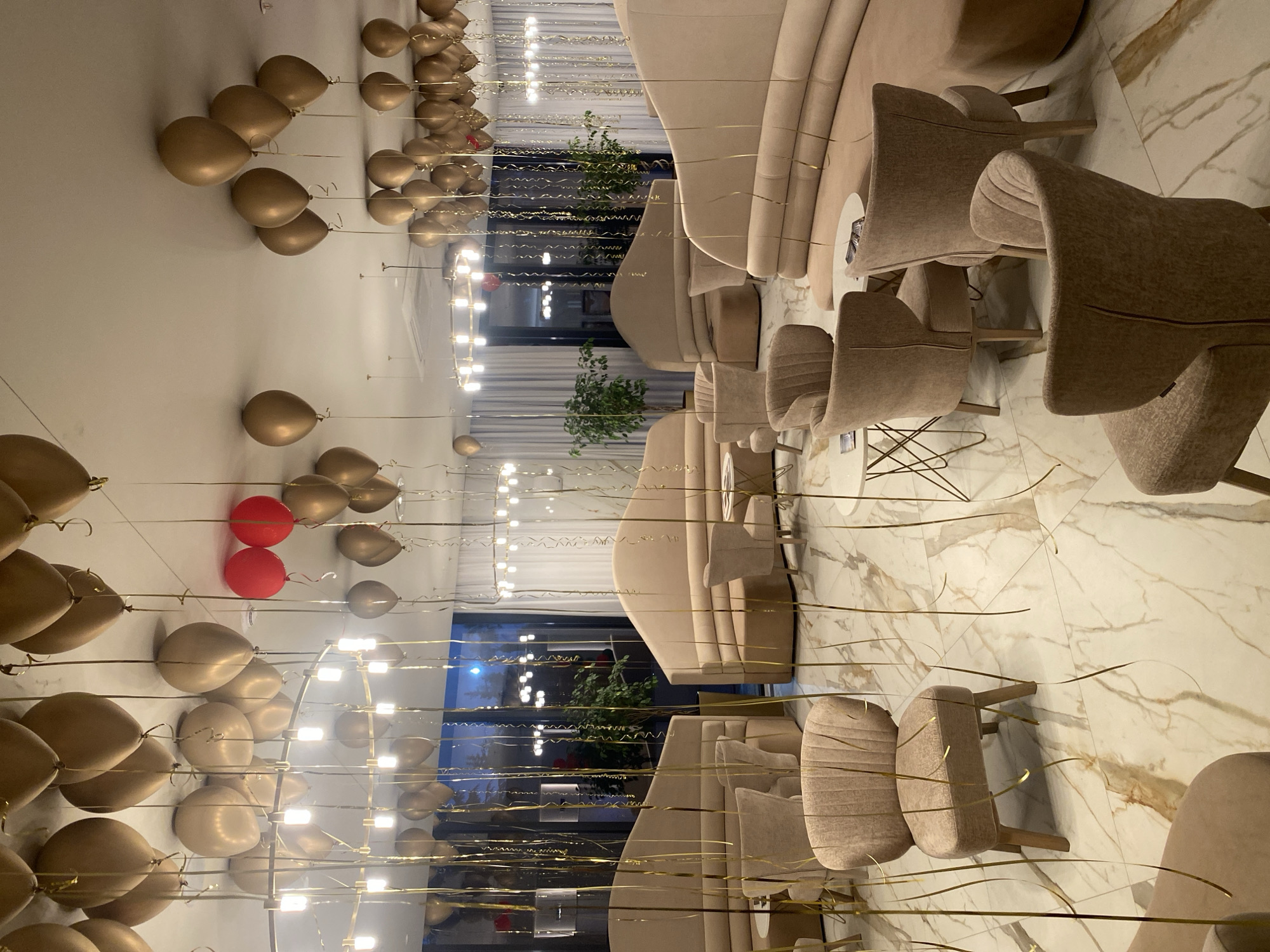 The image shows an indoor setting that appears to be a restaurant or event space, possibly located in the city of Sochi, Russia. The room is decorated with numerous gold balloons that are hanging from the ceiling. There are also two red balloons mixed in with the gold ones. The floor is a white marble with brown veining. The furniture consists of curved, light beige sofas and upholstered chairs. The room is well-lit, with a combination of natural light from windows and artificial light from chandeliers. There are plants visible near the windows. The overall impression is that the space is set up for a special occasion, such as a party or celebration. The reflection in the room seems to be of the same space and does not add any unique elements.