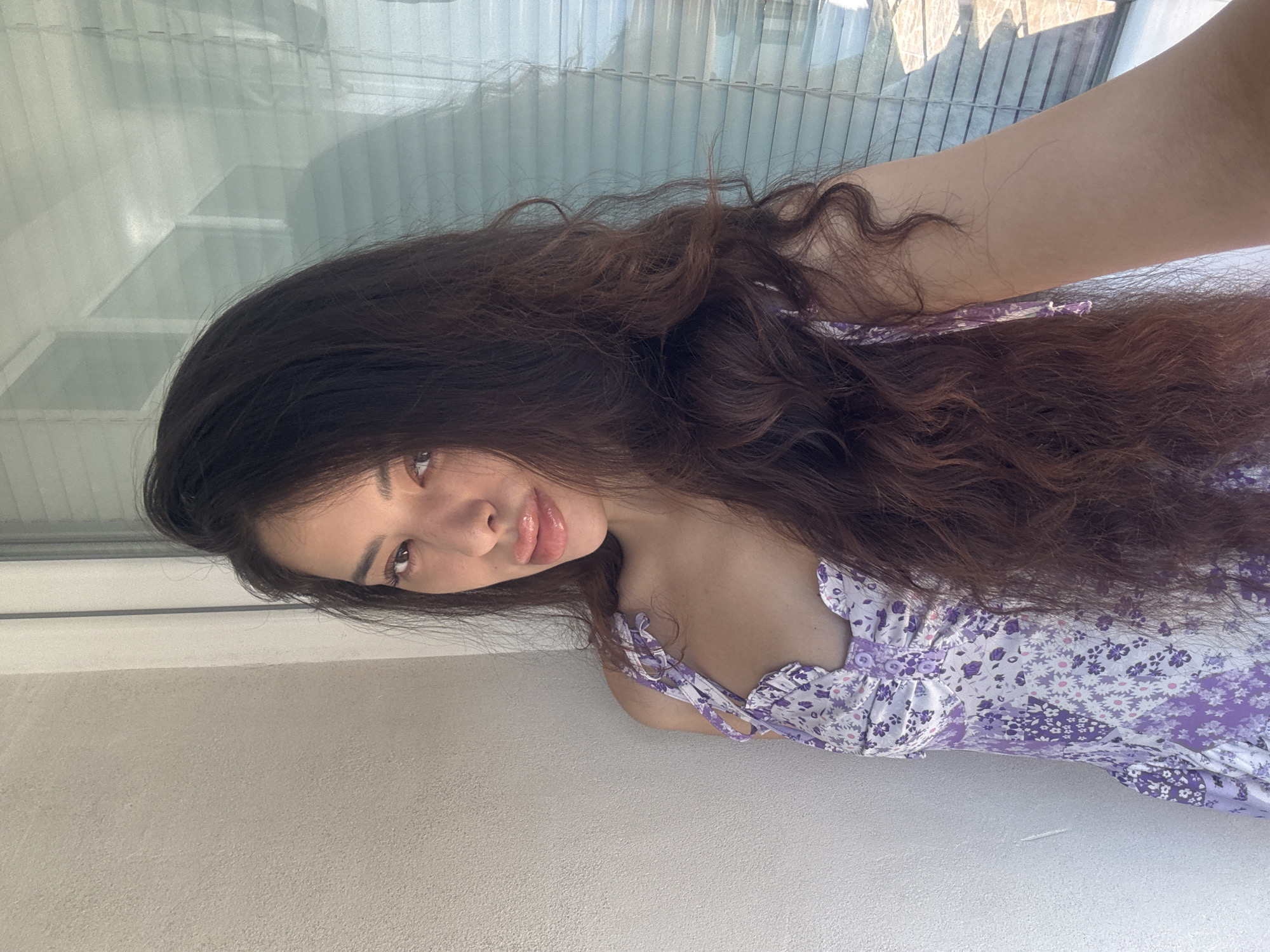 A woman with long, wavy brown hair and light skin is posing for a selfie indoors. She is wearing a light purple dress with a floral pattern. She is looking at the camera with a slight smile. Behind her is a window with closed blinds and a white wall below the window. The image does not provide enough information to determine the exact location in Plovdiv, Bulgaria.