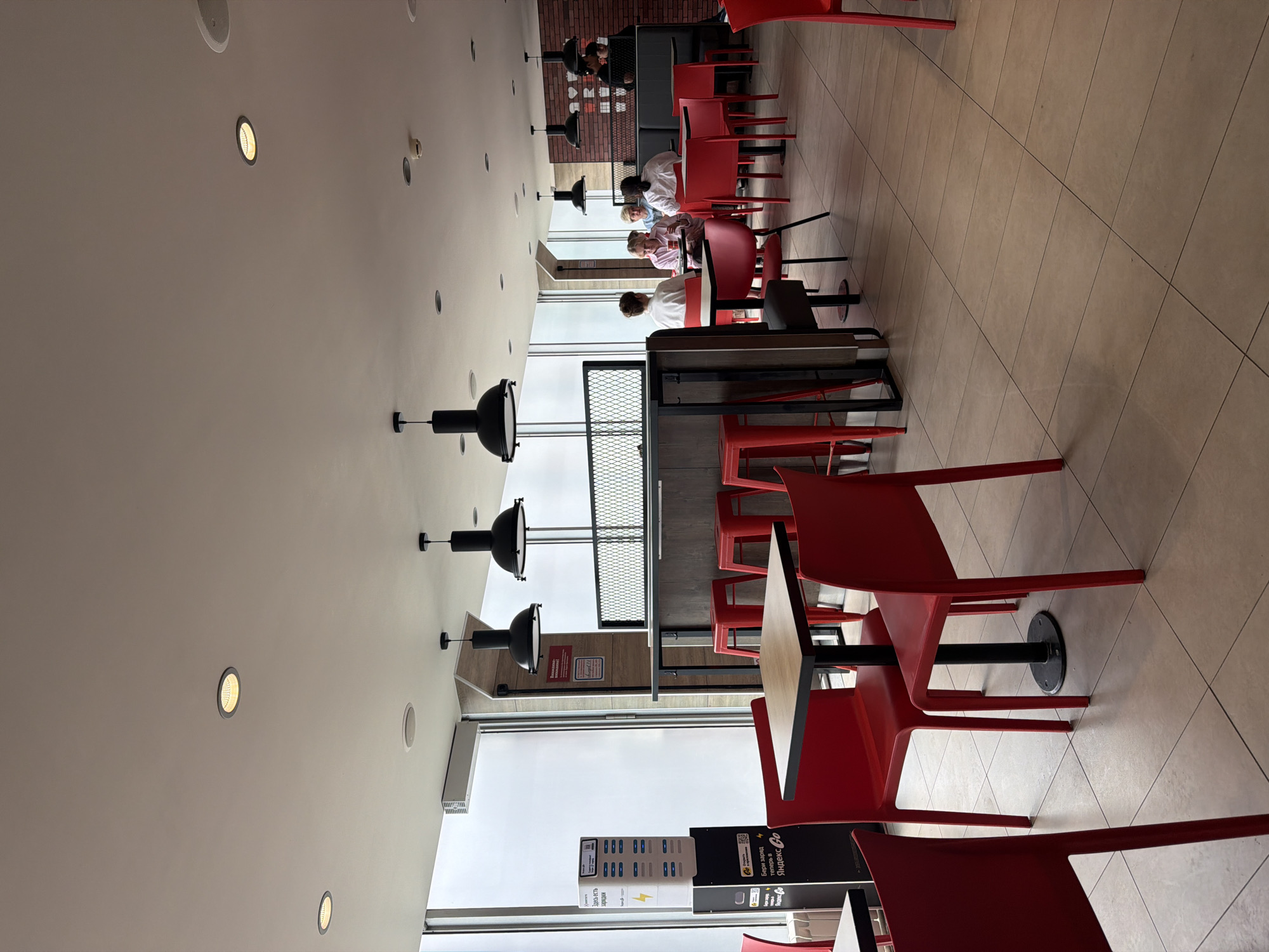 The image shows the interior of a fast-food restaurant in Moscow, Russia. The restaurant features rows of red chairs and tables. Several people are seated at the tables, some wearing white shirts. Pendant lamps hang from the ceiling. A charging station for electronic devices is located near a window. The floor is tiled.