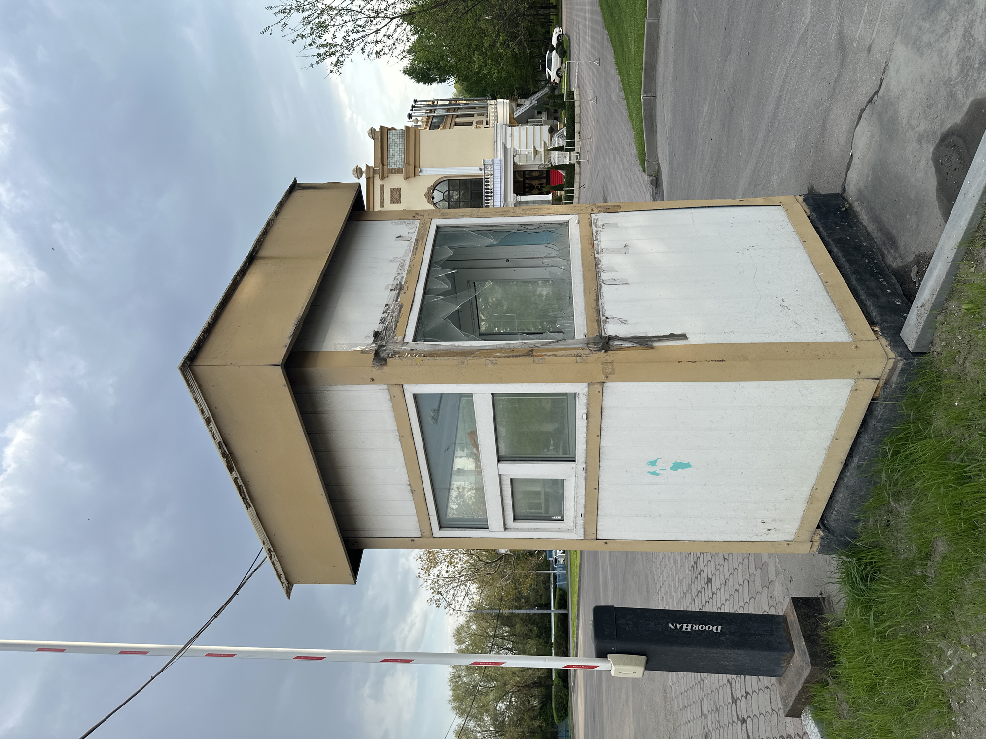 The image shows a dilapidated security booth located in Moscow, Russia. The booth has a white and beige exterior with two visible windows. The upper window is broken, with shattered glass evident. A lower window appears intact. A white and red striped barrier gate is partially visible in the foreground, attached to a black "DOORHAN" branded post. The booth is situated next to a paved area with grass growing nearby. In the background, a large, ornate building is visible, suggesting a relatively affluent or historically significant area. The sky is overcast.