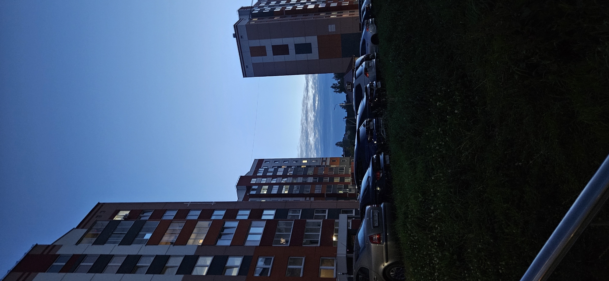 The image depicts a street scene in Vitsebsk, Belarus, at dusk. Two multi-story residential buildings dominate the background, with lights visible in some of the windows. In the foreground, a row of parked cars is lined up along a grassy area. The sky is a deep blue, transitioning into a lighter shade towards the horizon, with wispy clouds present. The overall lighting suggests the time is either early morning or late evening. There are no people or animals visible in the image, and no apparent activity is taking place.
