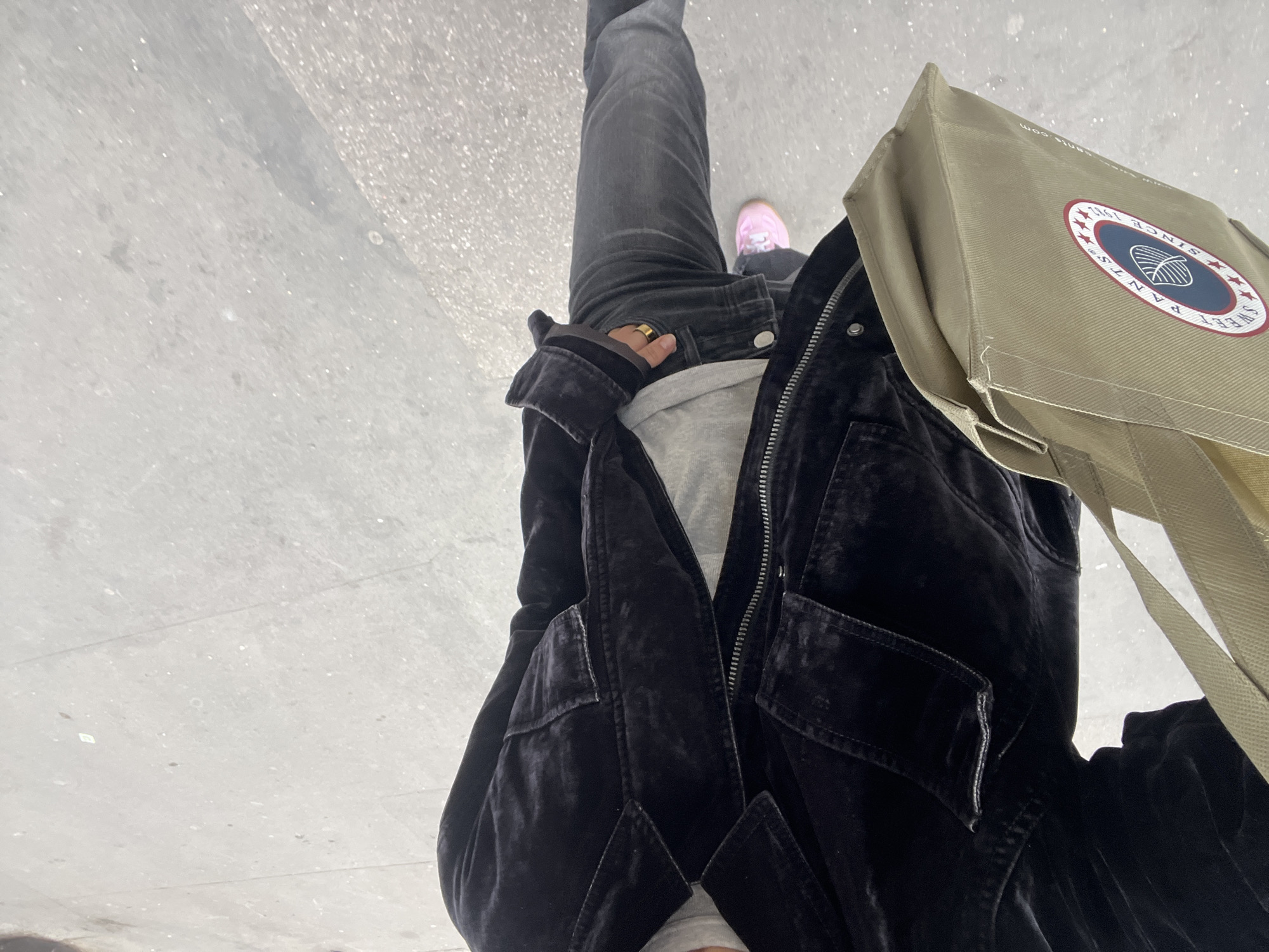 The image presents a high-angle, top-down view of a person from their mid-torso to their feet, standing on a light gray, textured ground that appears to be a concrete sidewalk or paved surface.
The person is wearing dark attire, including a black velvet jacket with a prominent zipper and large rectangular pockets on the front. A lighter gray t-shirt or sweatshirt is visible underneath the open jacket. Dark wash denim jeans cover the legs, and a gold-colored ring is worn on a finger of one hand, which is resting near the hip. The person is wearing light pink sneakers.
A khaki or tan canvas tote bag is slung over the shoulder, positioned on the right side of the frame. The bag features a circular logo with a red outer ring and a blue inner circle. White text in the blue circle clearly spells out "SWEET PANTS," and "SINCE 1961" is visible in white text on the red outer ring. A white graphic, resembling a yarn ball or spool, is centered within the blue circle. The top edge of the bag also shows partial text, including ".com," suggesting a website address.
The lighting indicates daytime, with soft, even illumination and no strong shadows, possibly suggesting an overcast sky. The scene offers an urban setting. While no specific landmarks are visible in the image, the provided context indicates the location is Paris, France.