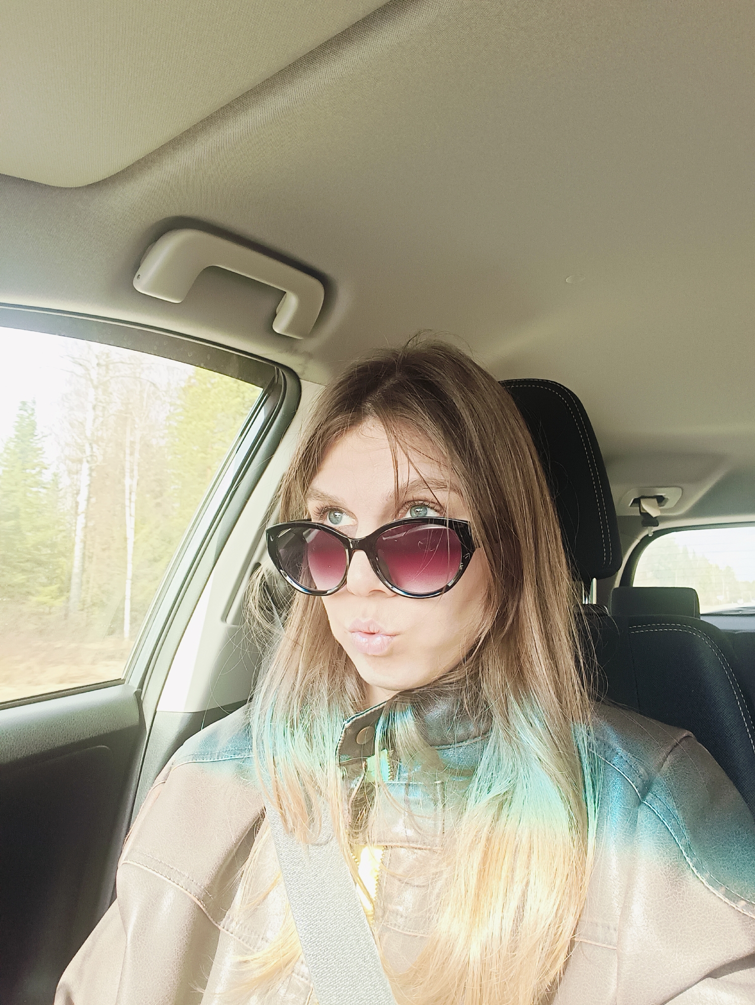 A young woman is depicted in a medium shot, seated in the passenger seat of a car during the daytime. She has long, light brown hair, which shows a subtle blue/green tint, likely from a light refraction effect. She is wearing cat-eye style sunglasses with dark, gradient lenses that transition to a reddish-purple. Her lips are pursed in a pouting or kissing expression, and her gaze is directed upwards and slightly to the right. She wears a light brown or beige leather-like jacket and has a seatbelt fastened across her chest.
The car's interior is visible, including the light-colored headliner, a grab handle, a black headrest with white stitching, and the window frame. Outside the car window, a bright, possibly sunny, environment with trees, some appearing like birches with light bark, and a hint of a road suggest a rural or natural setting. No visible text is present in the image.