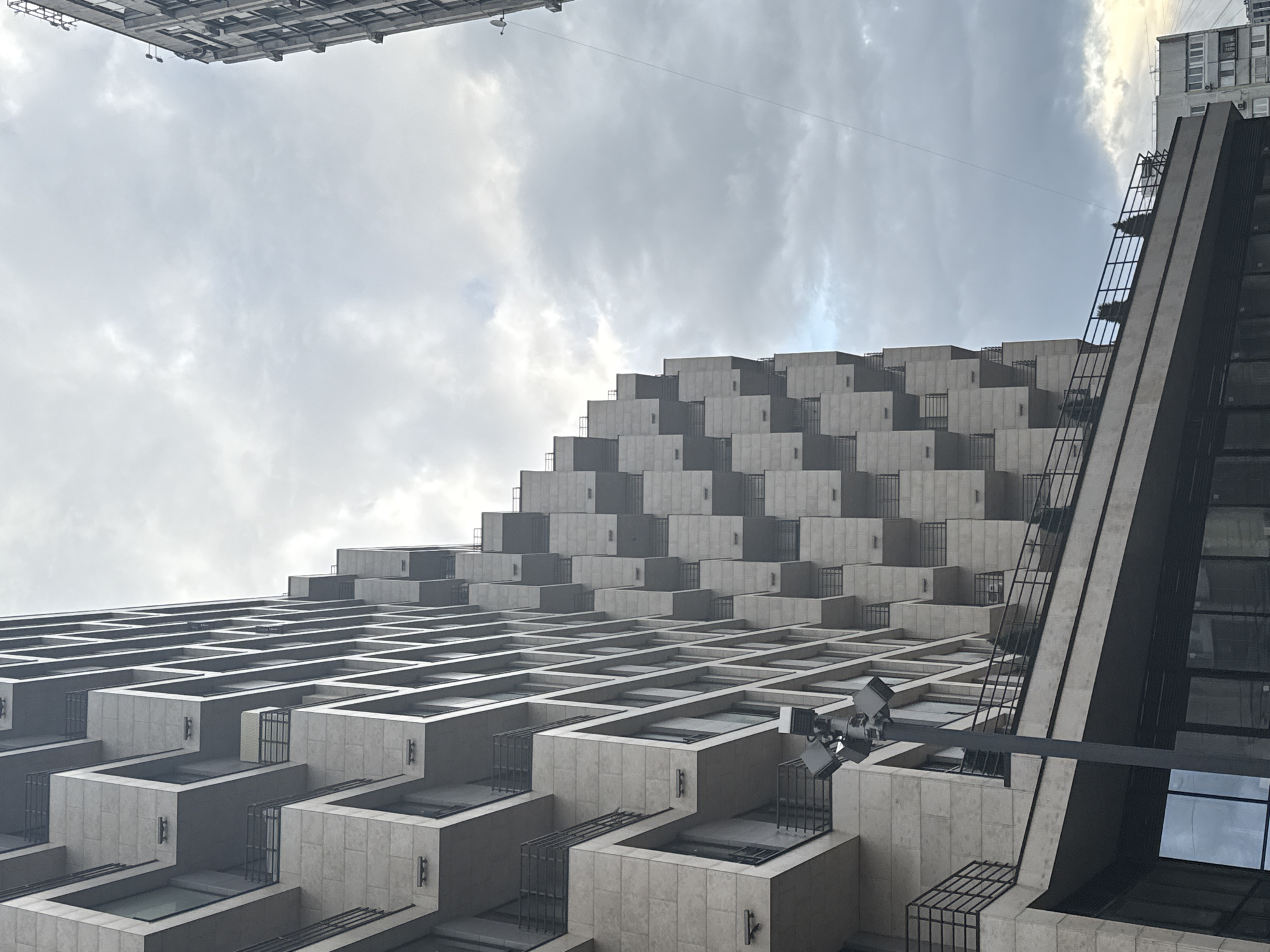 The photograph depicts a low-angle view of an architectural structure in Moscow, Russia, specifically the Dominion Tower. The building exhibits a modernist design with a stepped, terraced facade composed of cube-like modules. Each module features a recessed area that appears to function as a balcony or patio.
The structural elements are primarily constructed of light-colored concrete or stone, with black metal railings and window frames accentuating the geometric design. The sky is overcast, casting a soft, diffused light across the scene. Another building is visible at the top of the frame. A surveillance camera is mounted on one of the cube-like modules.