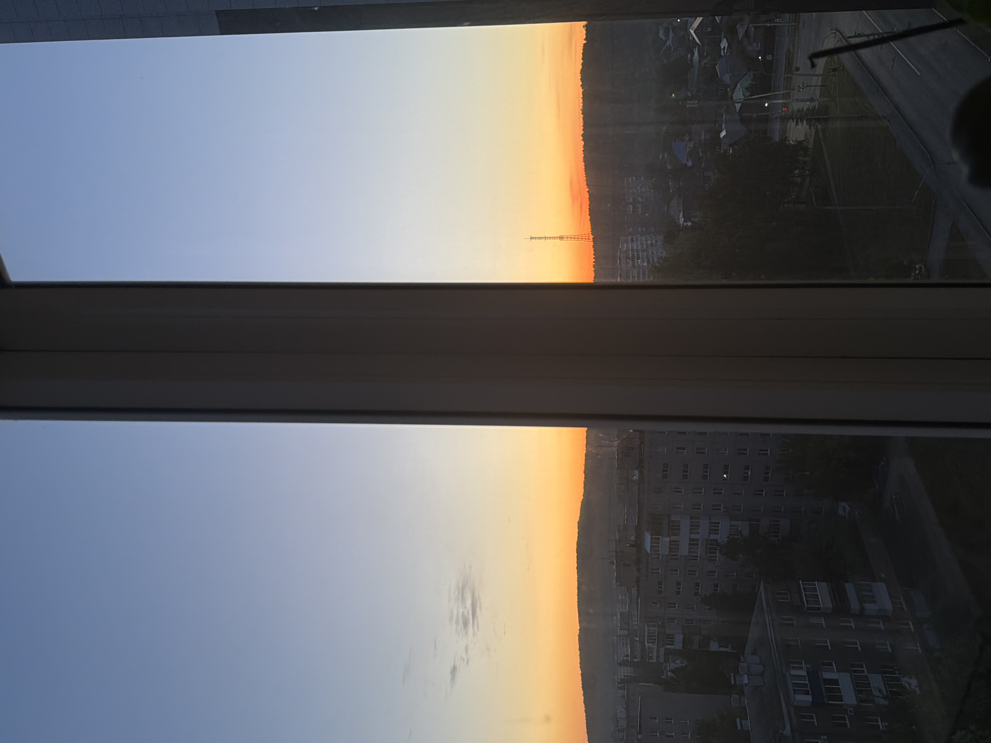 A window frames a view of a city at sunset. The sky transitions from blue at the top to orange near the horizon, which is lined with trees and buildings. A tall communication tower is visible against the sunset sky. Buildings and trees are reflected in the glass of the window. A road is also visible.