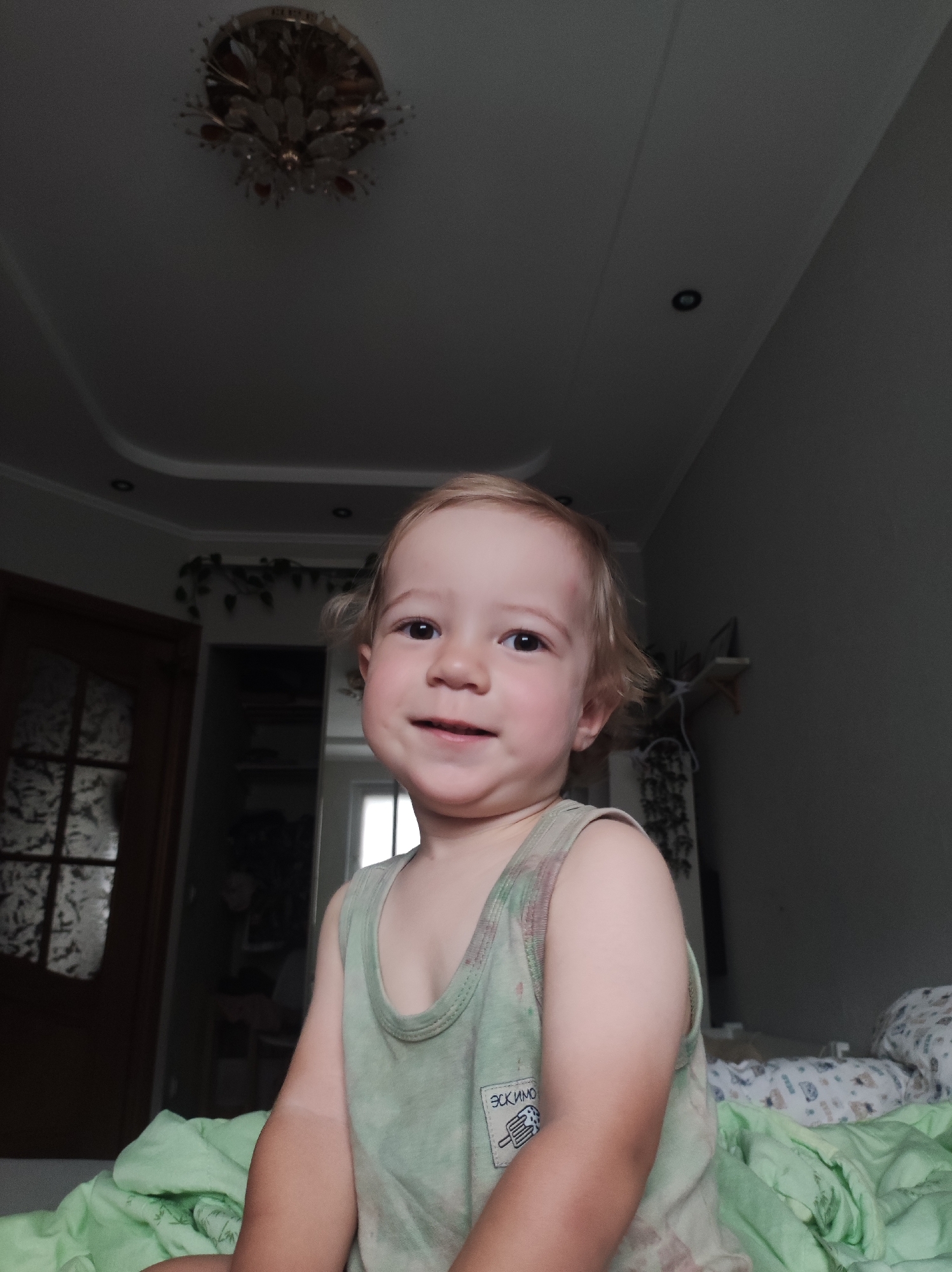 A close-up shot captures a young child, likely a toddler, sitting on a bed with light green bedding. The child has fair hair and is wearing a light green sleeveless shirt with a tag that has some text and an image of an ice cream cone. The child is smiling and looking directly at the camera. In the background, there's a door with frosted glass panels on the left, and on the right, a wall with a shelf holding various items. A ceiling light fixture is visible at the top of the frame. The overall lighting suggests the image was taken indoors, possibly in a bedroom. The location is a residential interior in the city of Chaikovsky, Russia.