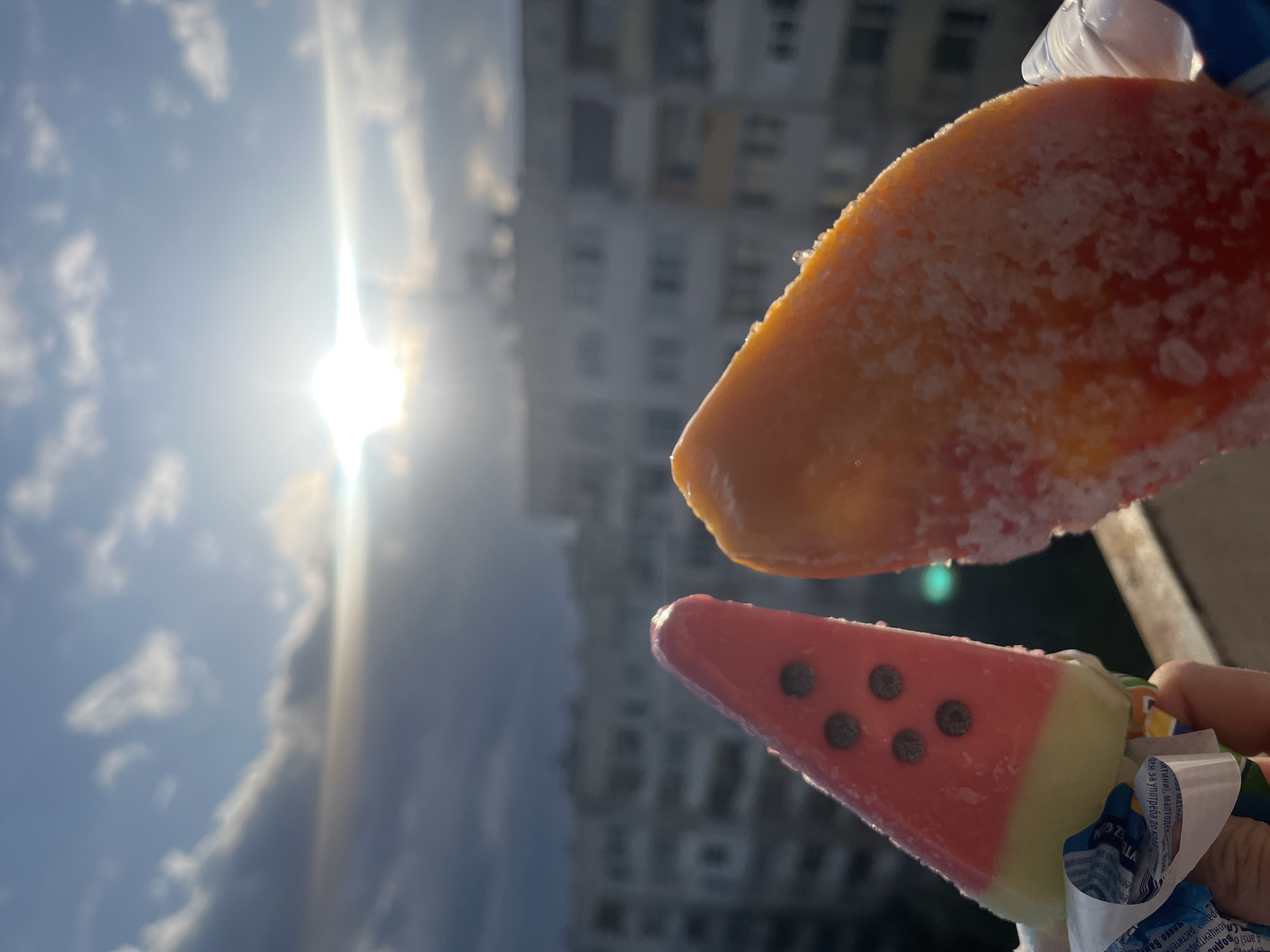 Here is a description of the image:
The photo shows two ice pops held up against a bright, sunny sky. The sun's glare is intense. A building, possibly an apartment block, is visible in the background, blurred by the depth of field. One ice pop is orange, potentially flavored with mango or citrus. The other is designed to look like a slice of watermelon, with a pink body decorated with chocolate chips to mimic seeds and a light green rind. The ice pops appear frosty, suggesting they have just been removed from the freezer. Part of a hand holding the ice pops, and some packaging are visible at the lower right corner of the image. Based on the text on the packaging, it's likely that the location is Sofia, Bulgaria.