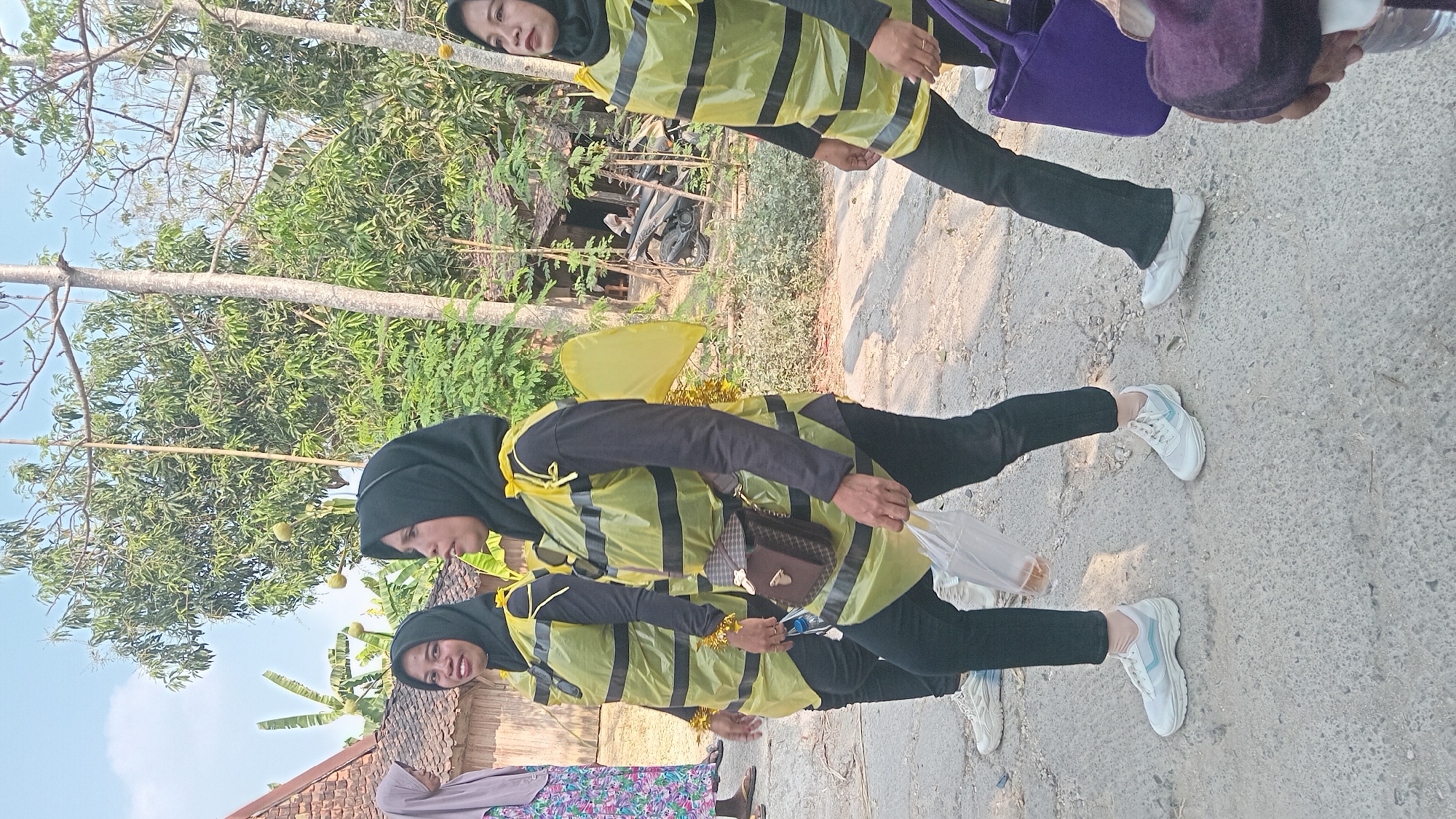 The image depicts three women walking outdoors in what appears to be a rural or suburban area of Kedungwaru, Indonesia. The women are dressed in matching costumes that resemble bee outfits, characterized by yellow vests with black stripes and what seem to be yellow wings attached to the back of two of the vests. They are wearing black hijabs and black pants. The woman on the left is wearing white sneakers, while the middle woman is wearing black pants and white sneakers. The woman on the right is wearing black pants and white sneakers, and is carrying a purple bag. One of the women is holding a plastic bag with bread inside. The background shows trees, vegetation, and a traditional-looking building with a tiled roof. The ground is paved with a rough, gray material, suggesting a road or pathway. The lighting indicates it is daytime, likely with bright sunlight.