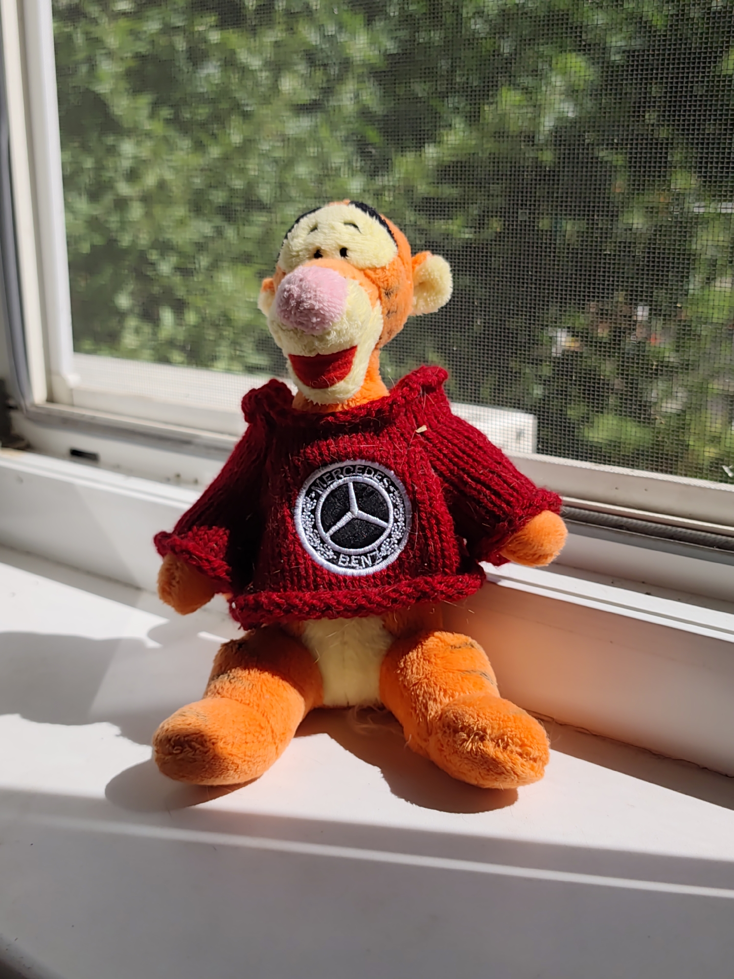 A stuffed toy resembling the Disney character Tigger is positioned on a white windowsill, facing the camera. The toy is wearing a red knitted sweater with a circular patch on the chest featuring the Mercedes-Benz logo. Behind the toy, a window screen is visible, obscuring a background of green foliage, suggesting an outdoor environment. The lighting indicates a sunny day, casting shadows on the windowsill. The toy is seated with its legs extended forward. The setting appears to be indoors, near a window. There is no indication of this location being in the city of Dzerzhinsk, Russia.