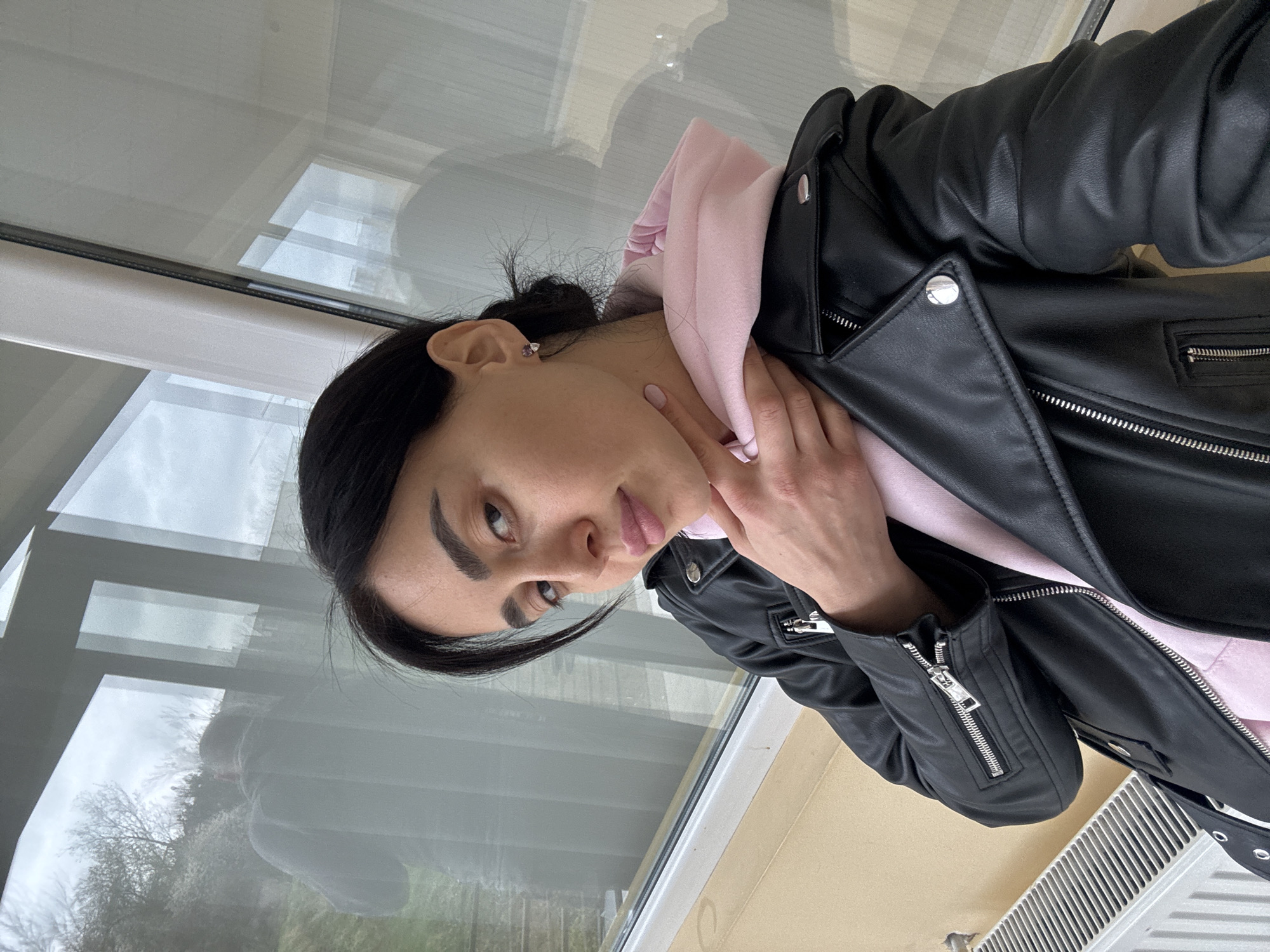 A woman is standing indoors near a window. She is wearing a black leather jacket over a pink hooded sweatshirt. She has dark hair pulled back in a bun and is wearing stud earrings. Her right hand is lightly touching her neck. Outside the window, trees and other vegetation are visible in an overcast sky. A radiator is partially visible in the bottom right corner. Based on the architectural style of the window and the surrounding vegetation, it is plausible that the photo was taken in Tbilisi, Georgia, but more detailed features would be needed for confirmation.