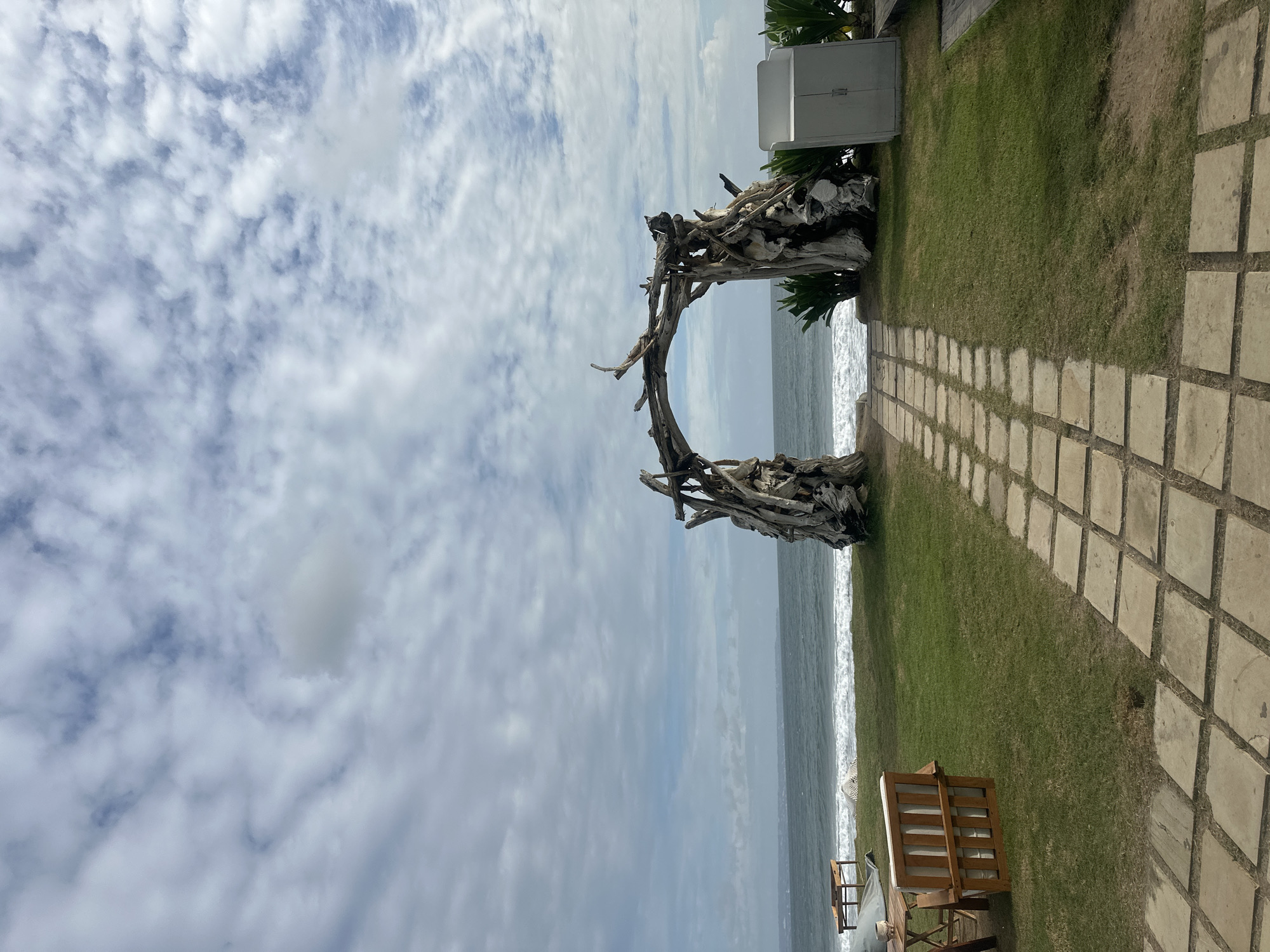 The image shows a beach scene in North Kuta, Indonesia. In the foreground, a stone-paved pathway leads to a grassy area, where a circular driftwood sculpture stands. Beyond the sculpture is the ocean with white waves. The sky is cloudy. Adjacent to the pathway on the right is a wooden bench. Adjacent to the sculpture on the left, is a white cabinet.