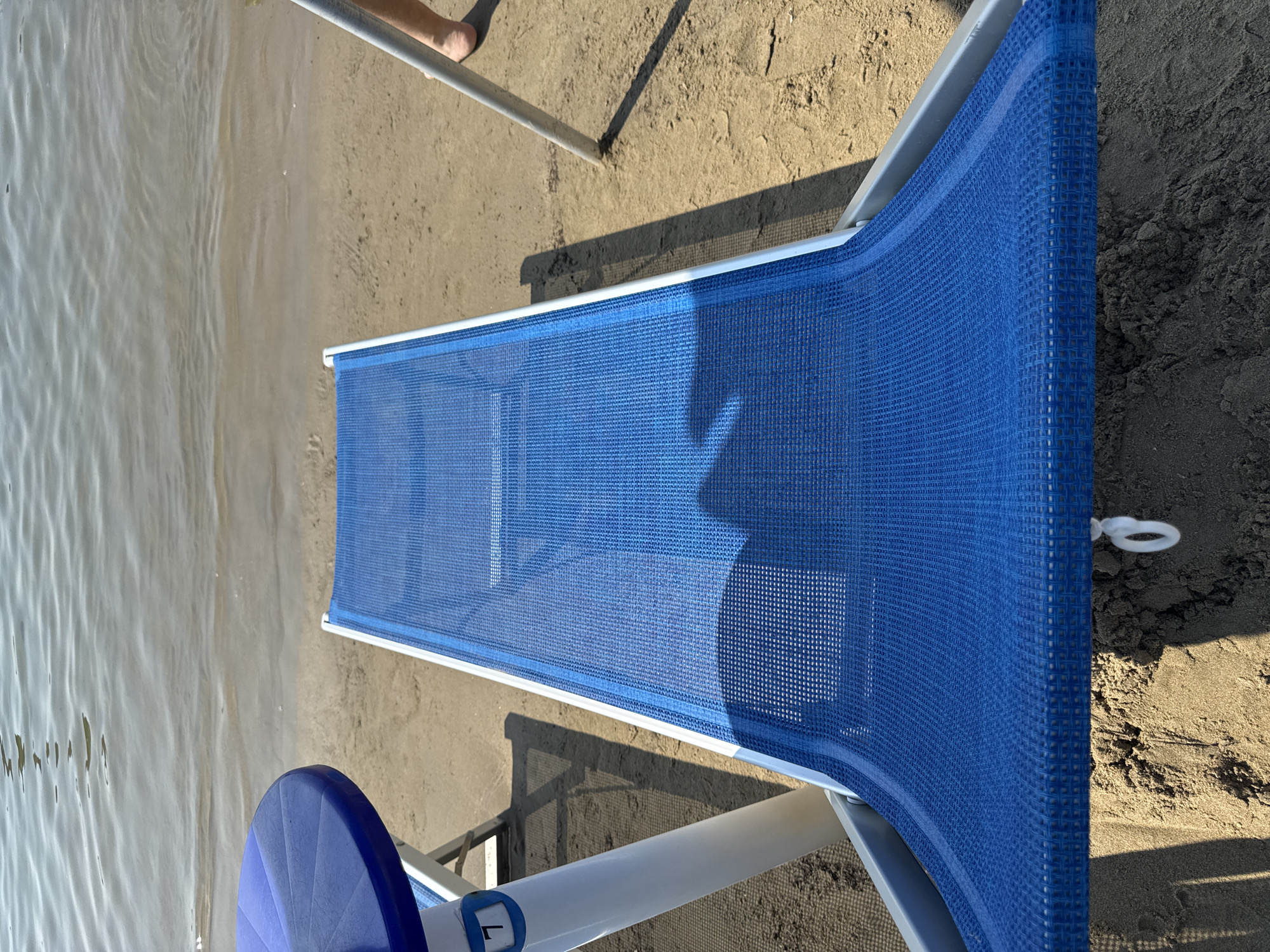 The image depicts a close-up, elevated view of a blue mesh beach lounger positioned on a sandy shore, with the gentle waves of a body of water visible in the background. The lounger is centered in the frame, and its textured surface, made of small, repeating squares, is clearly visible. To the left of the lounger, a portion of a blue circular table top is visible, supported by white poles. A blue marker with the number '7' is attached to one of the white poles. To the right of the lounger, a thin metal pole is partially embedded in the sand, and a person's bare leg and foot are visible behind it, suggesting someone is sitting or standing nearby. The sand is a light brown color, with some darker, wetter sand closer to the water's edge. Shadows are cast across the sand, indicating a sunny day. The water exhibits subtle ripples and a light brown hue, consistent with shallow coastal or lake water. The scene is set on a beach, likely in the Liguria region of Italy, given the mention of Sanremo.