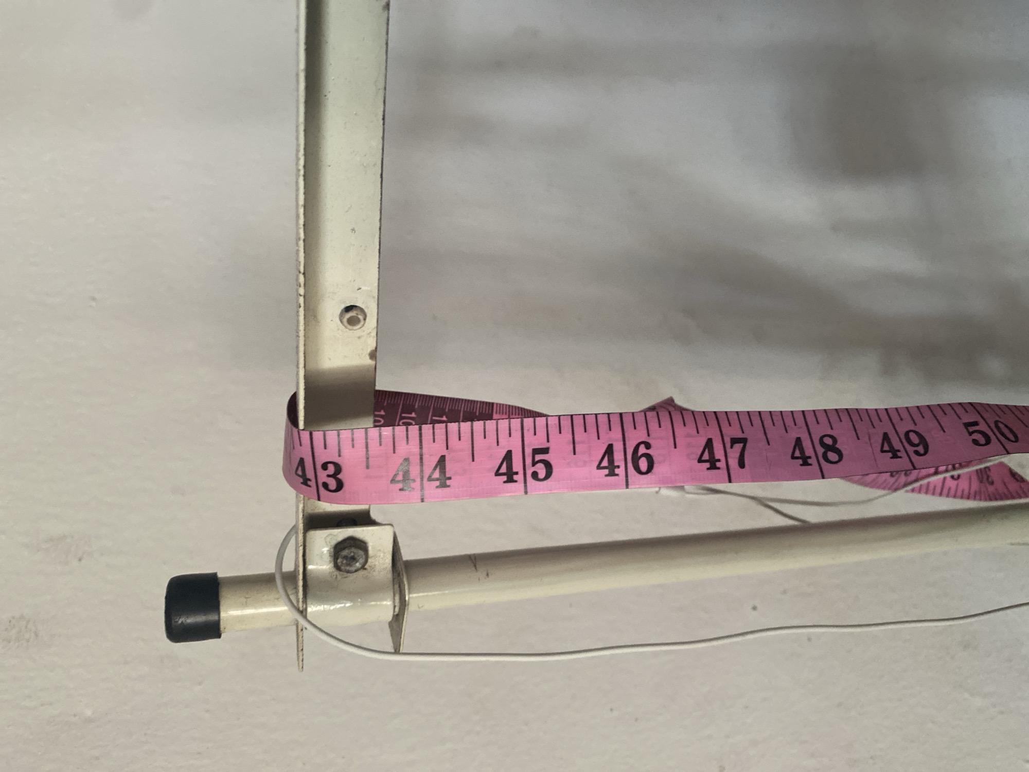 The image shows a close-up of a pink measuring tape hanging from a cream-colored metal frame. The measuring tape has black markings and numbers, with visible measurements ranging from 43 to 50. The metal frame is attached to a vertical cream-colored pole with a black cap at the top. A white wire is also visible, attached to the frame and hanging down. The background is a textured, off-white wall. There is no visible indication of the city of Jalingo, Nigeria, in the image.