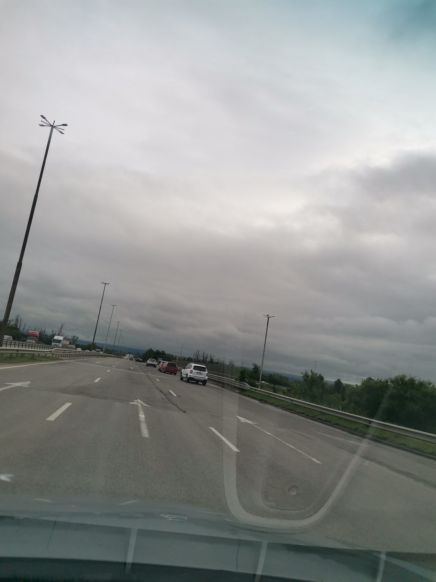 An overcast day is depicted on a highway in an unknown location. The highway has multiple lanes, with traffic moving in the same direction. Several vehicles, including cars and trucks, are visible on the road. Tall streetlights are positioned along the side of the highway. The sky is filled with gray clouds, suggesting a potentially rainy or gloomy atmosphere. Green trees and vegetation line the edges of the highway, and a metal guardrail separates the lanes from the embankment. The image appears to be taken from inside a vehicle, as the lower portion of the frame shows a reflection on a windshield.