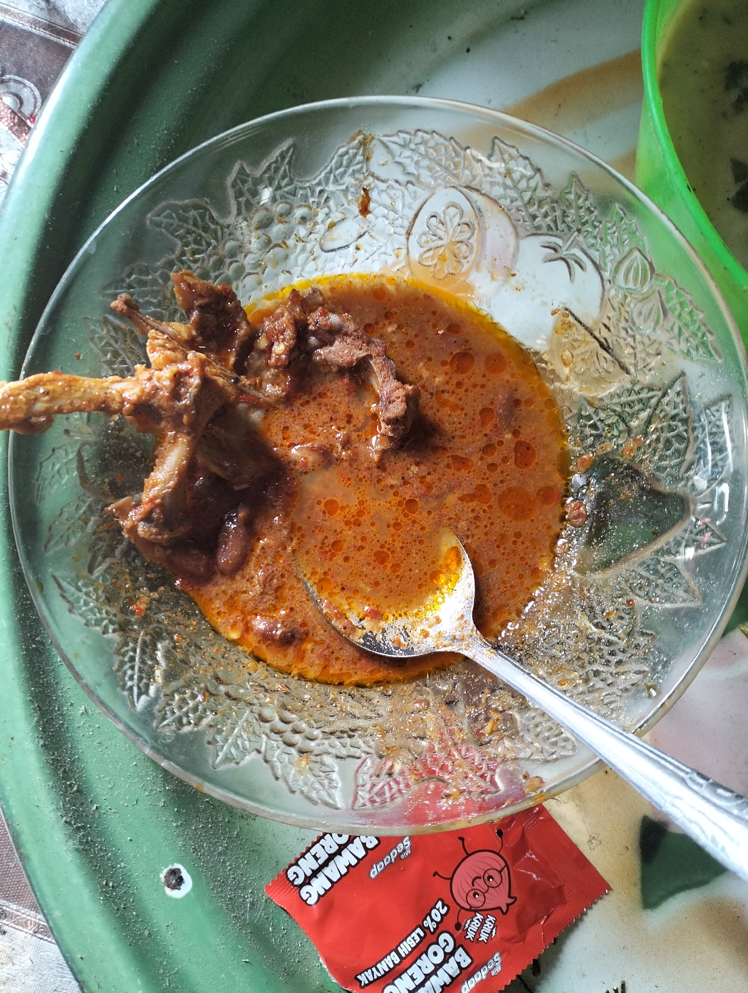 A close-up, top-down view shows a clear glass bowl containing a serving of meat in a rich, orange-red sauce. The meat appears to be bone-in pieces, possibly chicken or a similar fowl, coated in the flavorful sauce which has a slightly oily sheen and is dotted with small bubbles. A silver spoon rests partially submerged in the sauce, suggesting the meal is being eaten or has recently been consumed. The bowl itself has an ornate, patterned rim with embossed floral designs. It sits on a green, textured surface, likely a placemat or tray, which is itself on a patterned fabric surface. To the bottom right, a small, red sachet is visible, featuring an illustration of a cartoon onion wearing glasses and the text "SEDAP", and "BAWANG GORENG" and "20% LEBIH BANYAK", indicating it is likely a sachet of fried onion seasoning. The overall scene suggests a casual meal setting, potentially in a home or eatery. While the image itself does not provide explicit details about the city of Mandala Jaya, Indonesia, the presence of Indonesian text on the sachet strongly implies this location.