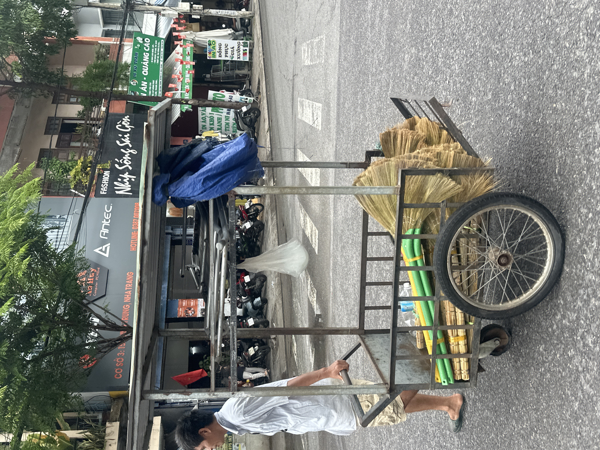 A man wearing a white shirt and tan shorts is pushing a cart filled with brooms and green plastic pipes on a street in Nha Trang Ward, Vietnam. The cart is metal-framed with two wheels. The sidewalk is visible in the background, lined with parked motorbikes and buildings featuring commercial signs. A sign for "Antec" and "Nhịp Sống Sai Gòn" is visible, along with signs advertising printing and advertising services.